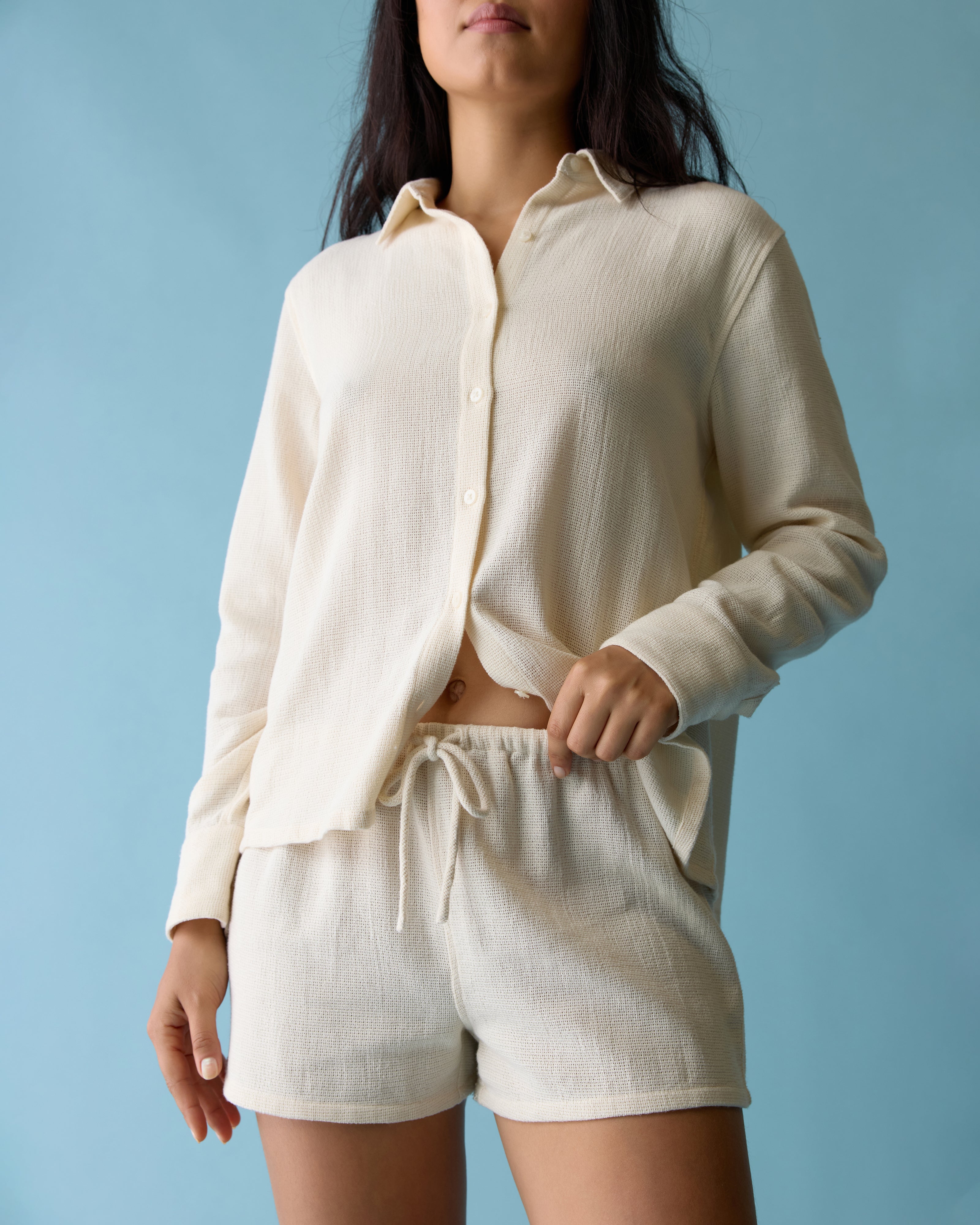 A woman stands against a blue background, wearing a cream-colored button-up shirt and matching Dandy Del Mar Sevilla Short - Scallop with an elastic waistband.