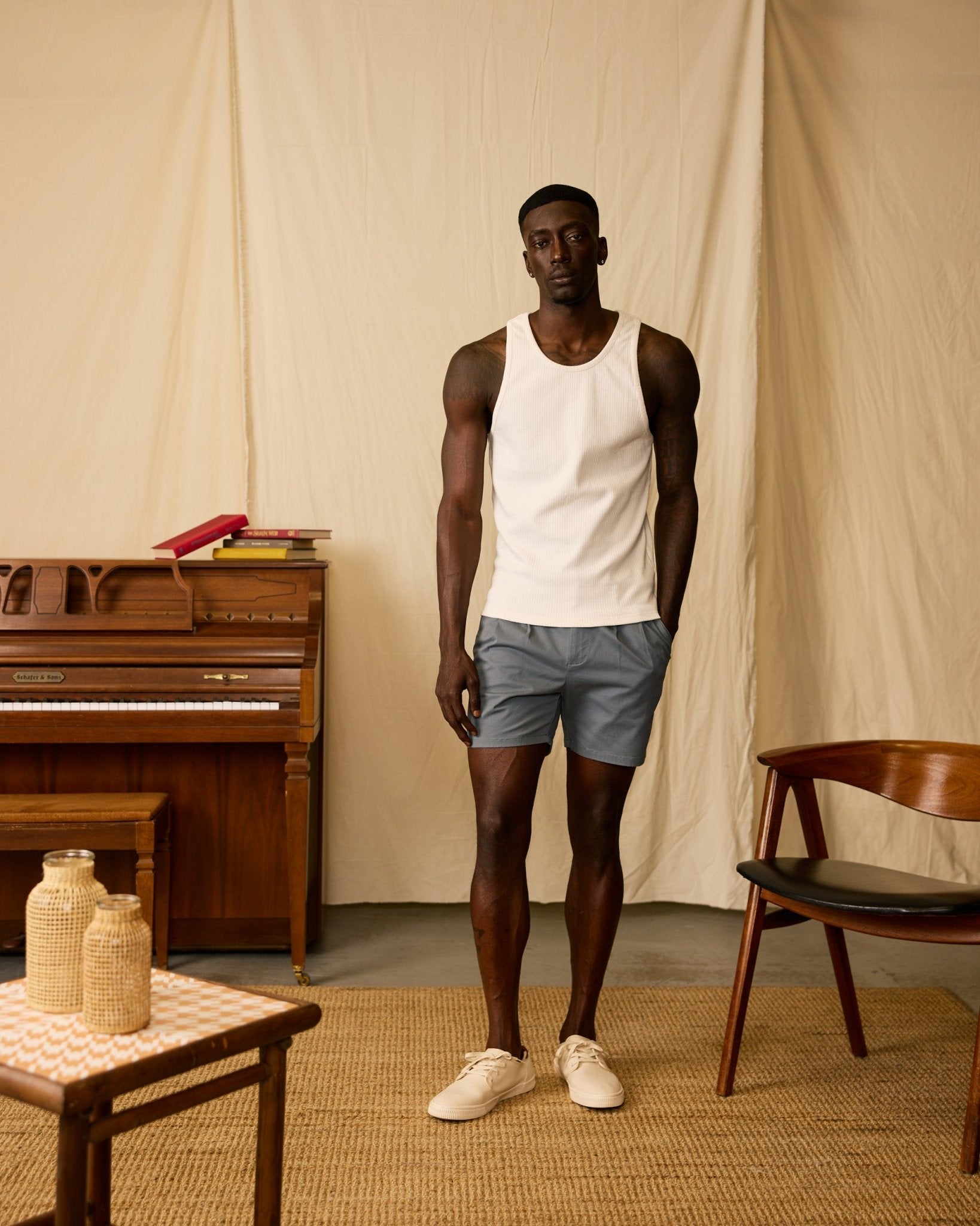 A man in a white tank top and Dandy Del Mar's The Rhodes Twill Shorts - Abyss stands indoors near a piano and wooden chair, surrounded by neutral décor.