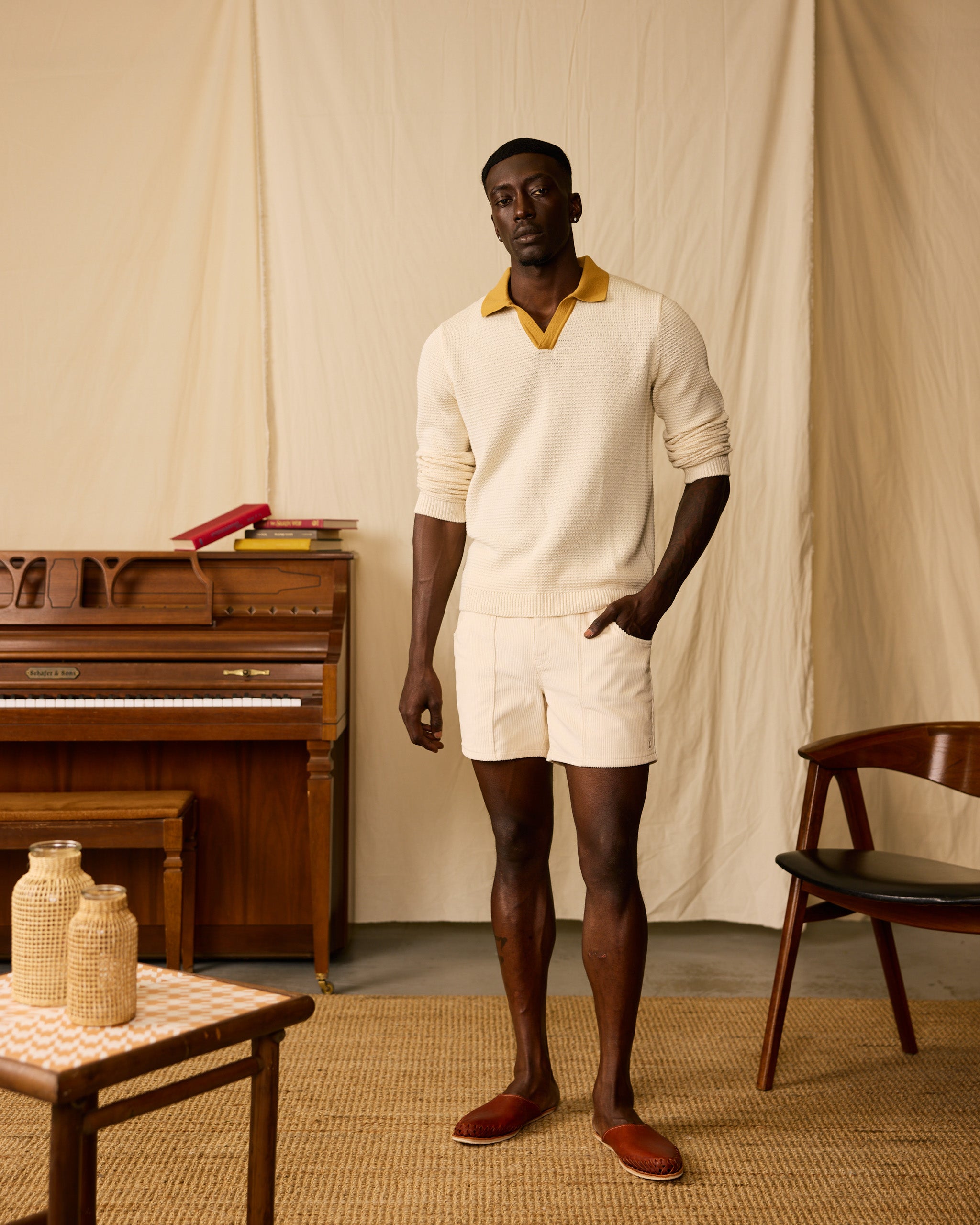 A man stands indoors in a cream knit sweater with a yellow collar, Dandy Del Mar’s The Corsica Corduroy Shorts - Scallop (5" inseam), and brown slip-on shoes. Behind him are a piano, books, chair, and table.