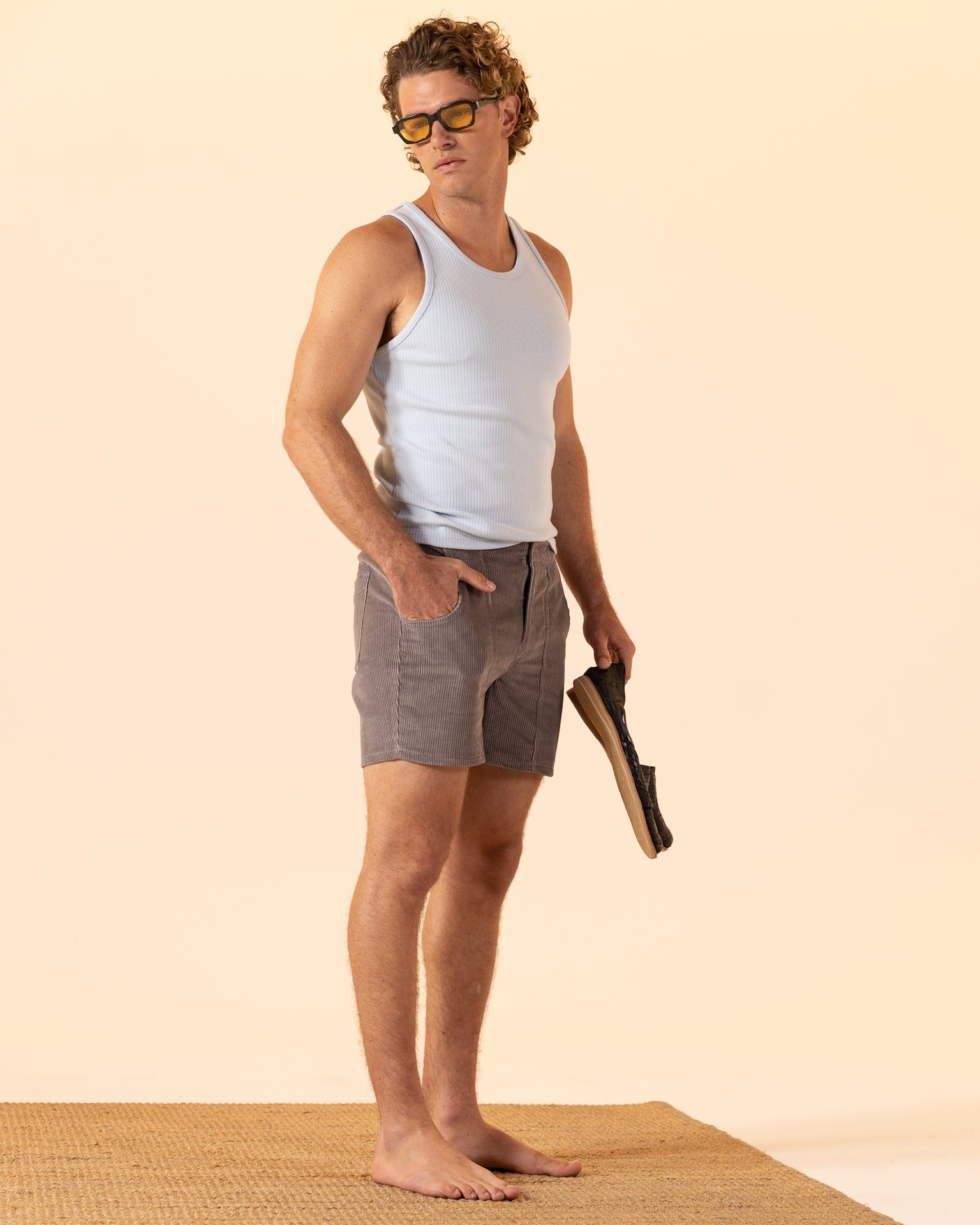 A man in The Milan Rib Tank - Comet by Dandy Del Mar and gray shorts stands barefoot on a woven mat against a beige background, holding a magazine and embodying luxury in simplicity.