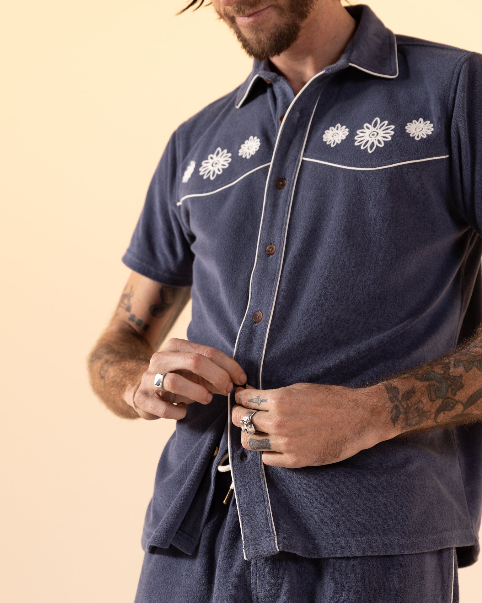 Embodying an Equatorial Cowboy style, this individual wears Dandy Del Mar's "The Gaucho Shirt - Moontide," a short-sleeve navy shirt with white floral embroidery. Buttoning up, their wrists and fingers showcase intricate rings and tattoos for rugged elegance.