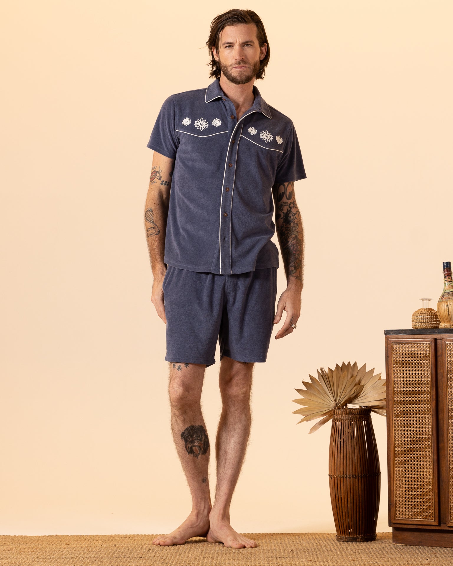 A tattooed man channels the Equatorial Cowboy look in Dandy Del Mar's Gaucho Shirt - Moontide with matching shorts, all made of terry cloth. He stands barefoot on a woven rug, surrounded by decorative pottery and a lush plant.
