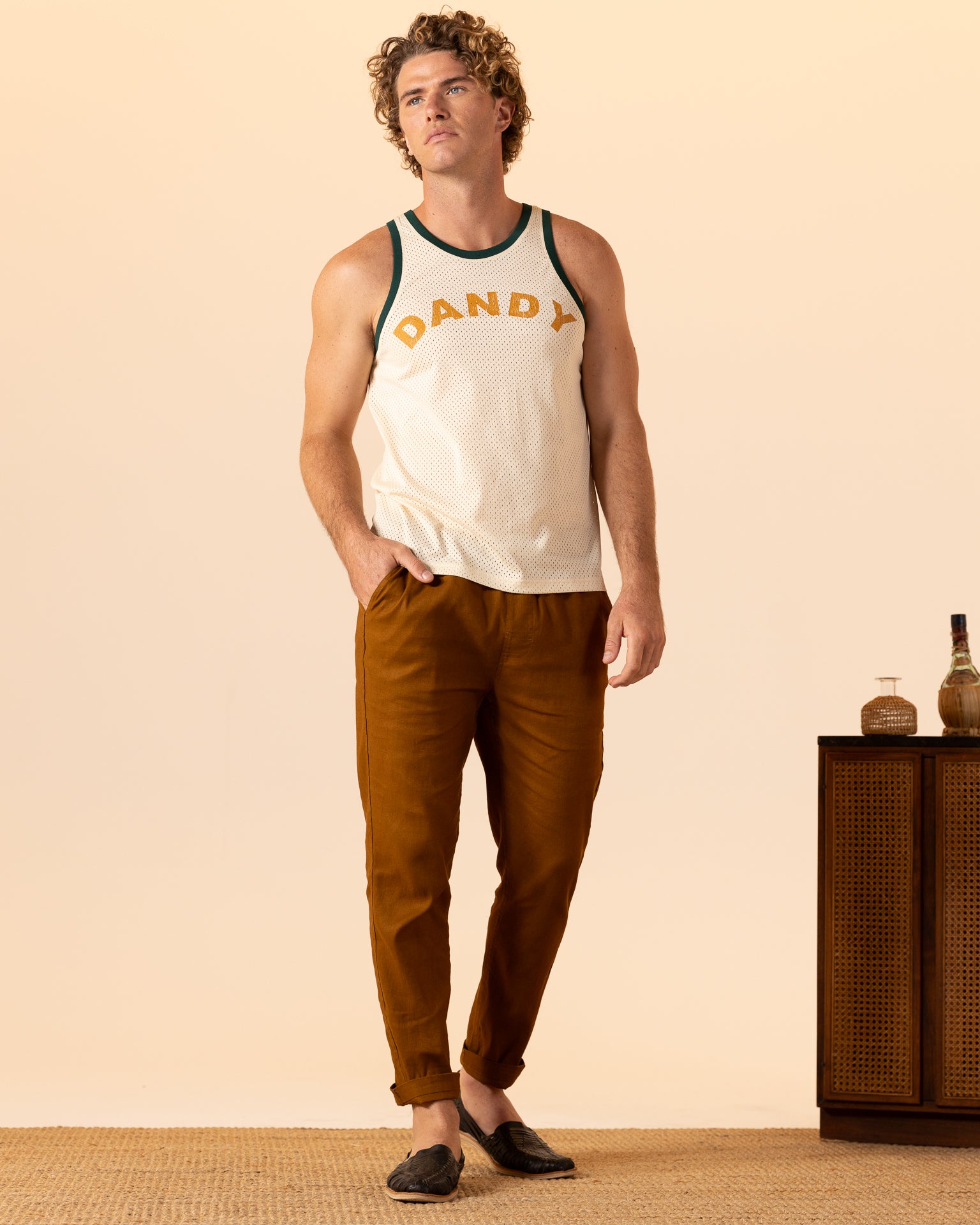 A person wearing The Kaena Mesh Tank - Sabbia by Dandy Del Mar, brown pants, and black shoes stands with one hand in pocket next to a wooden cabinet with bottles.