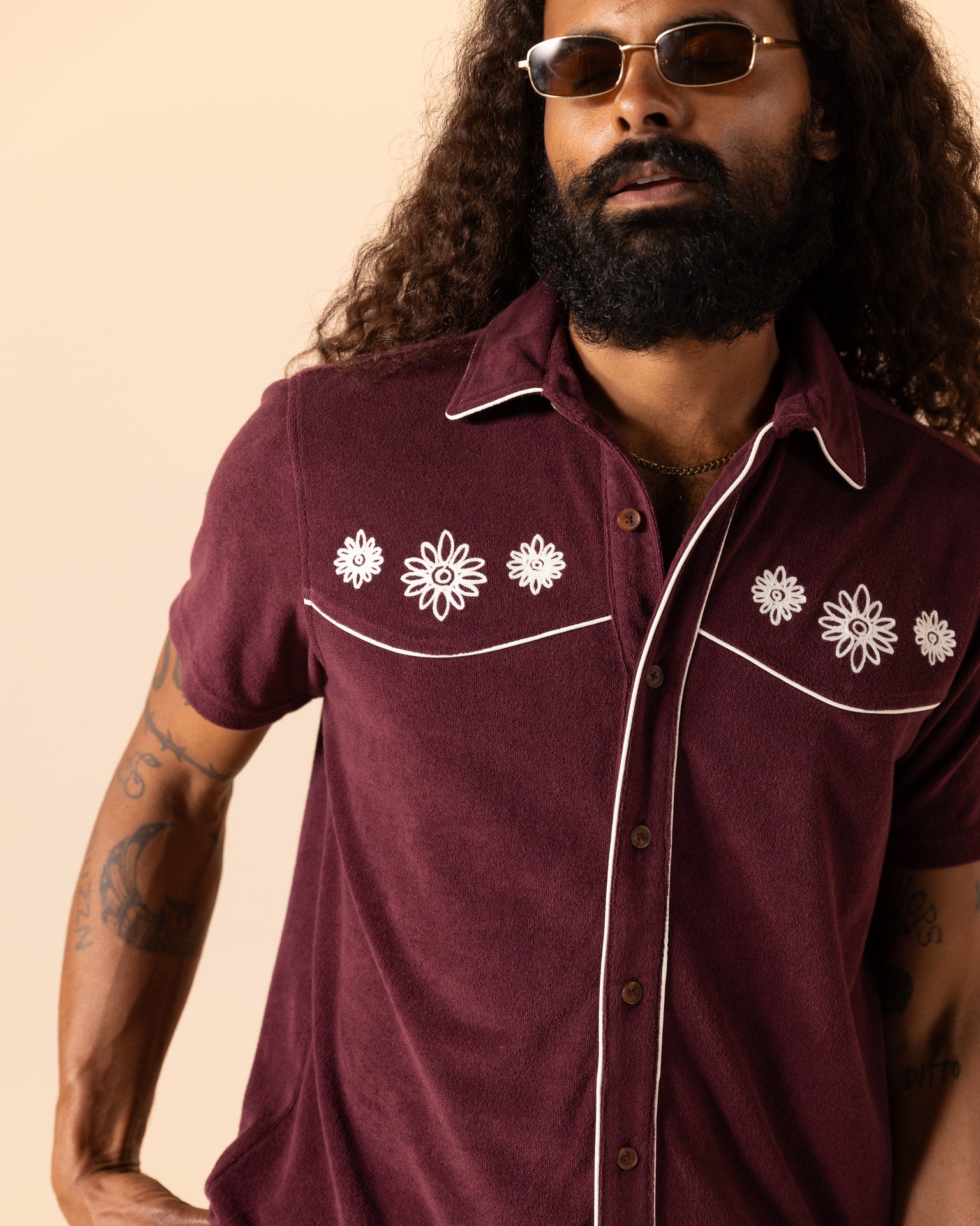 Sporting the Equatorial Cowboy style, a long-haired, bearded individual stands against a beige backdrop in sunglasses and Dandy Del Mar's The Gaucho Shirt - Bordeaux with floral embroidery.