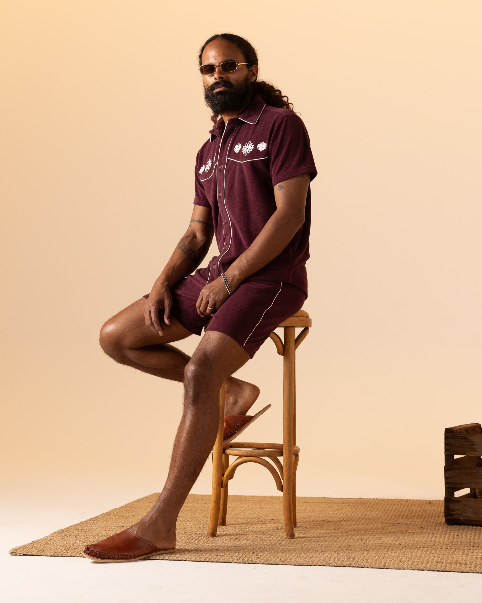 Sporting a full beard and long hair, a man wearing The Gaucho Short in Bordeaux by Dandy Del Mar sits on a wooden stool atop a woven rug, perfectly embodying the Equatorial Cowboy look.