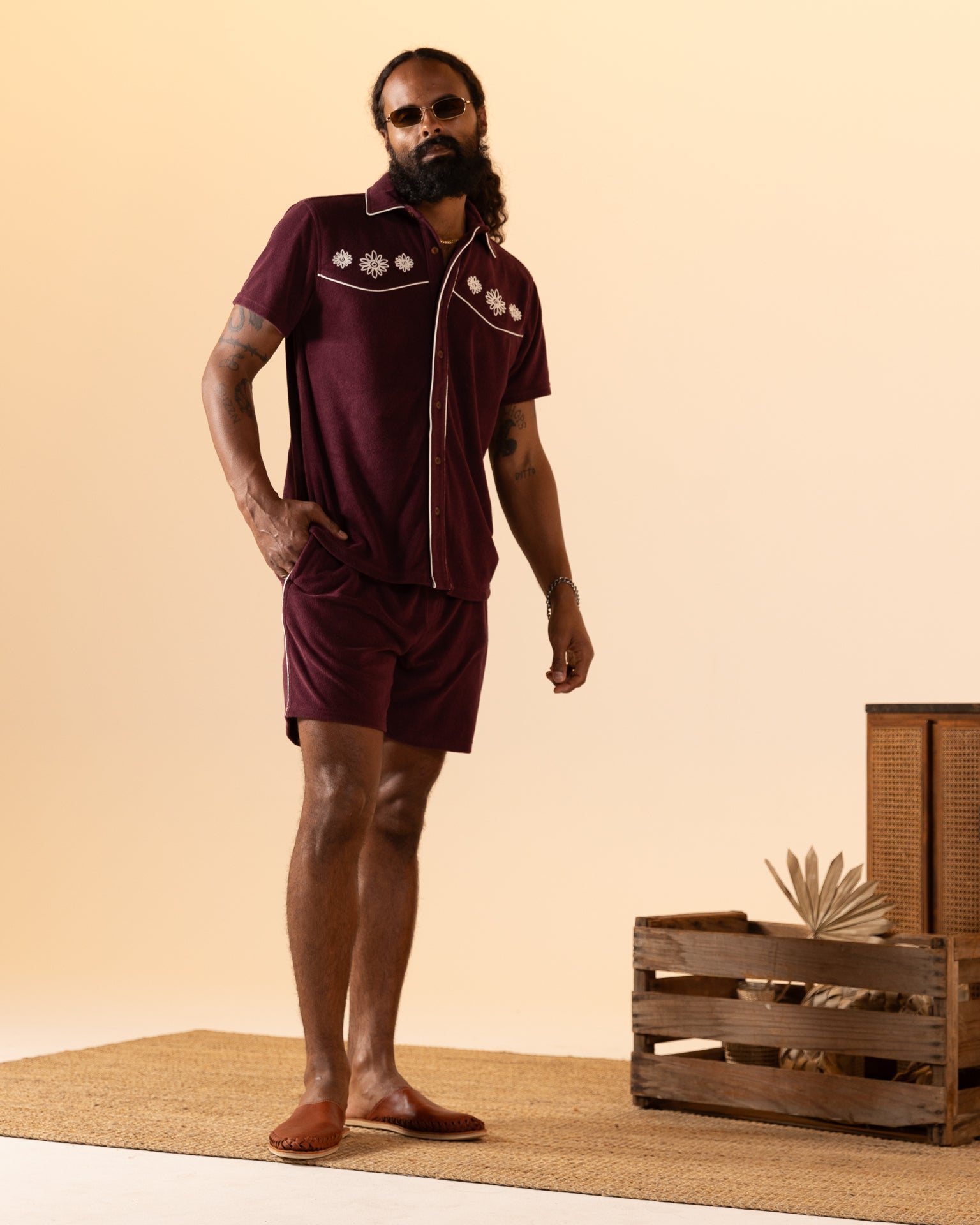 A man models The Gaucho Short - Bordeaux by Dandy Del Mar in a studio with a light brown backdrop. A wooden crate filled with lush plants adds an equatorial cowboy vibe to the scene.