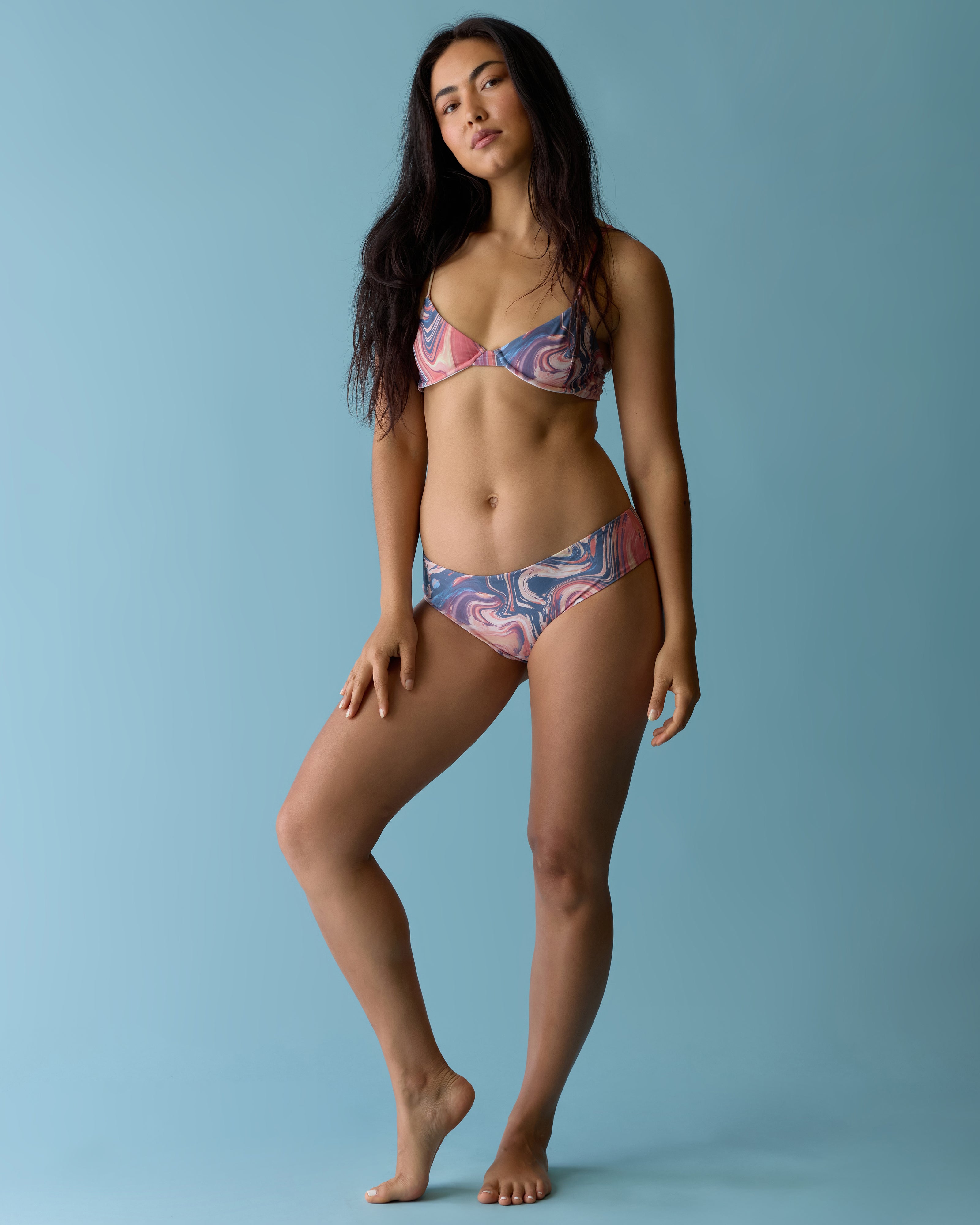 A woman stands barefoot against a light blue background, wearing Dandy Del Mar's The Avila Top - Lantana, an underwire bralette bikini made from recycled nylon. She has a neutral expression and relaxed posture.