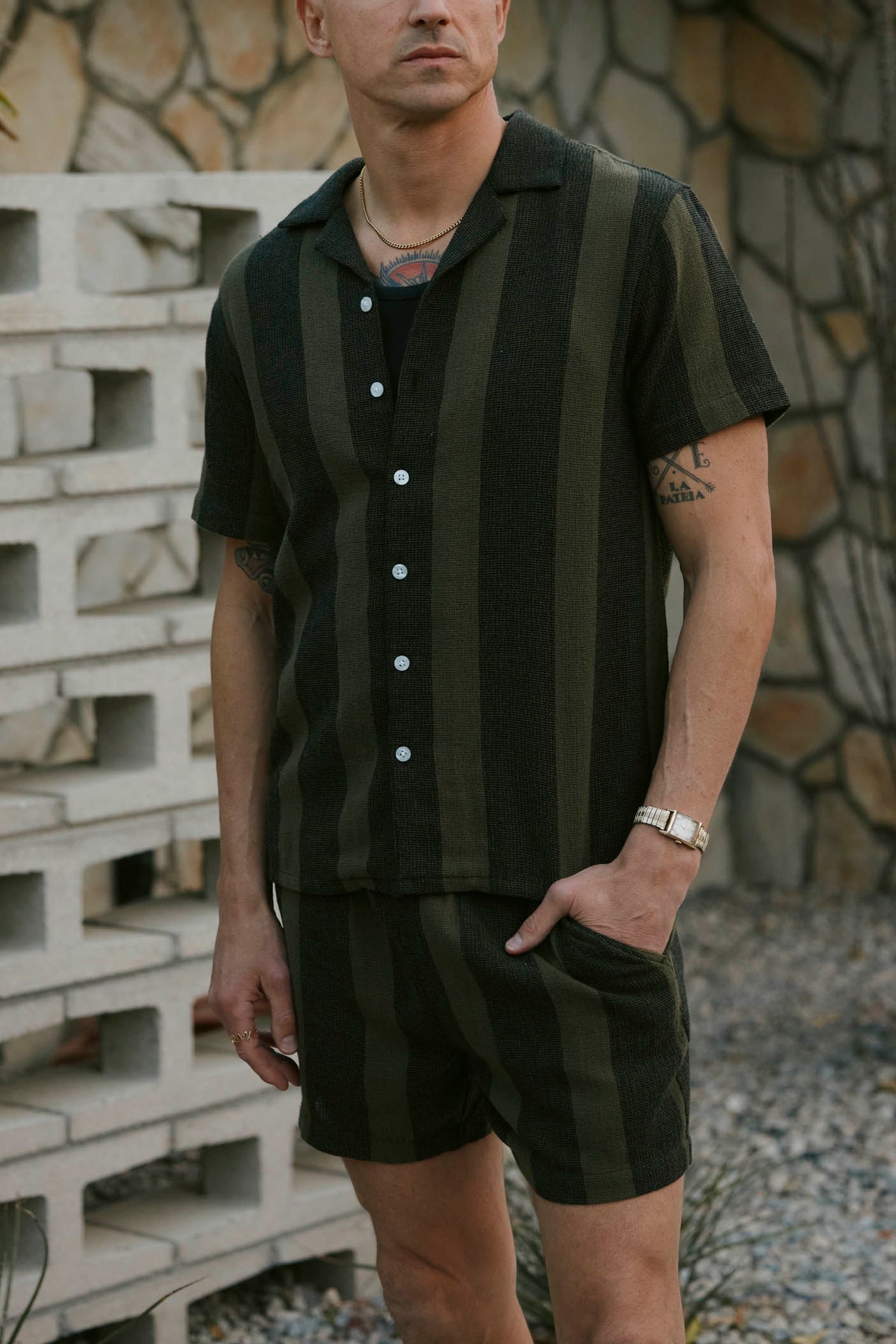 A man stands outside wearing Dandy Del Mar’s The Palma Shorts in Onyx, a relaxed fit short-sleeve, button-up and matching shorts set featuring dark green and black yarn dyed stripes, with one hand in his pocket.