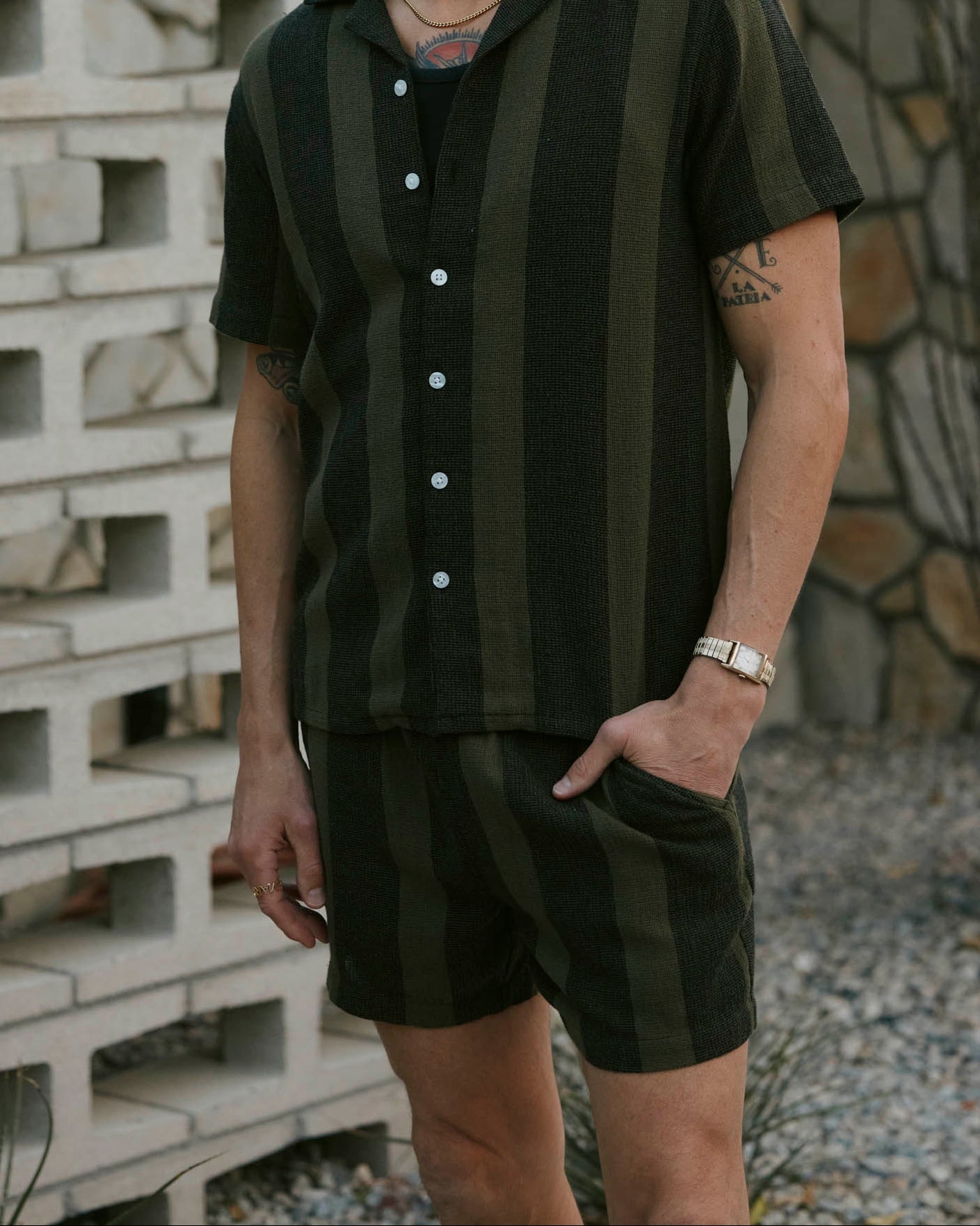 A man stands outside wearing Dandy Del Mar’s The Palma Shorts in Onyx, a relaxed fit short-sleeve, button-up and matching shorts set featuring dark green and black yarn dyed stripes, with one hand in his pocket.