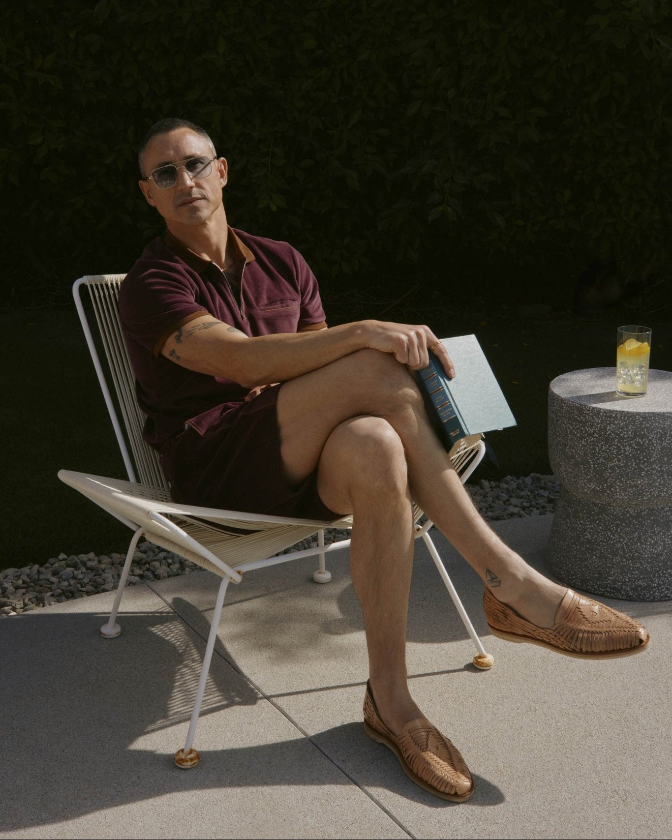 A person in Dandy Del Mar’s The Rio Velour Shorts in Bordeaux and sunglasses sits on a patio chair with legs crossed, holding a book. A drink rests on a nearby round stone table.
