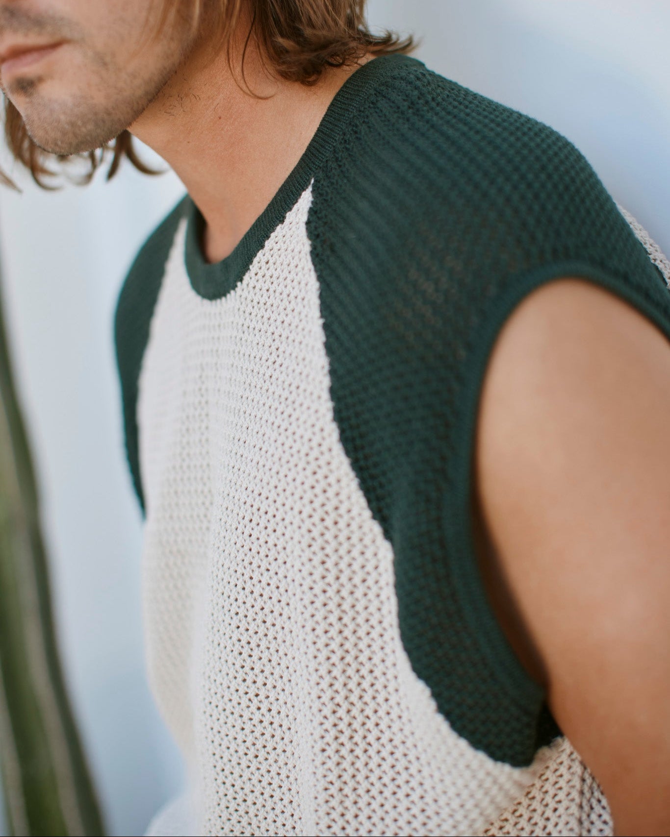 A person with shoulder-length brown hair wears The Porto Crochet Tank - Sabbia by Dandy Del Mar, a sleeveless cropped top with dark green trim and white mesh fabric, against a softly blurred background.
