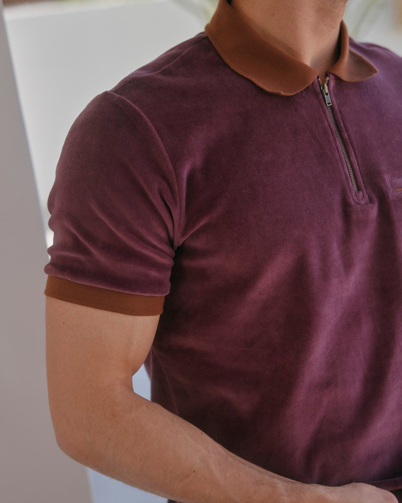 A man wears The Rio Velour Shirt - Bordeaux by Dandy Del Mar, a short-sleeve maroon polo with a brown collar and zipper, shown from shoulders to mid-torso.