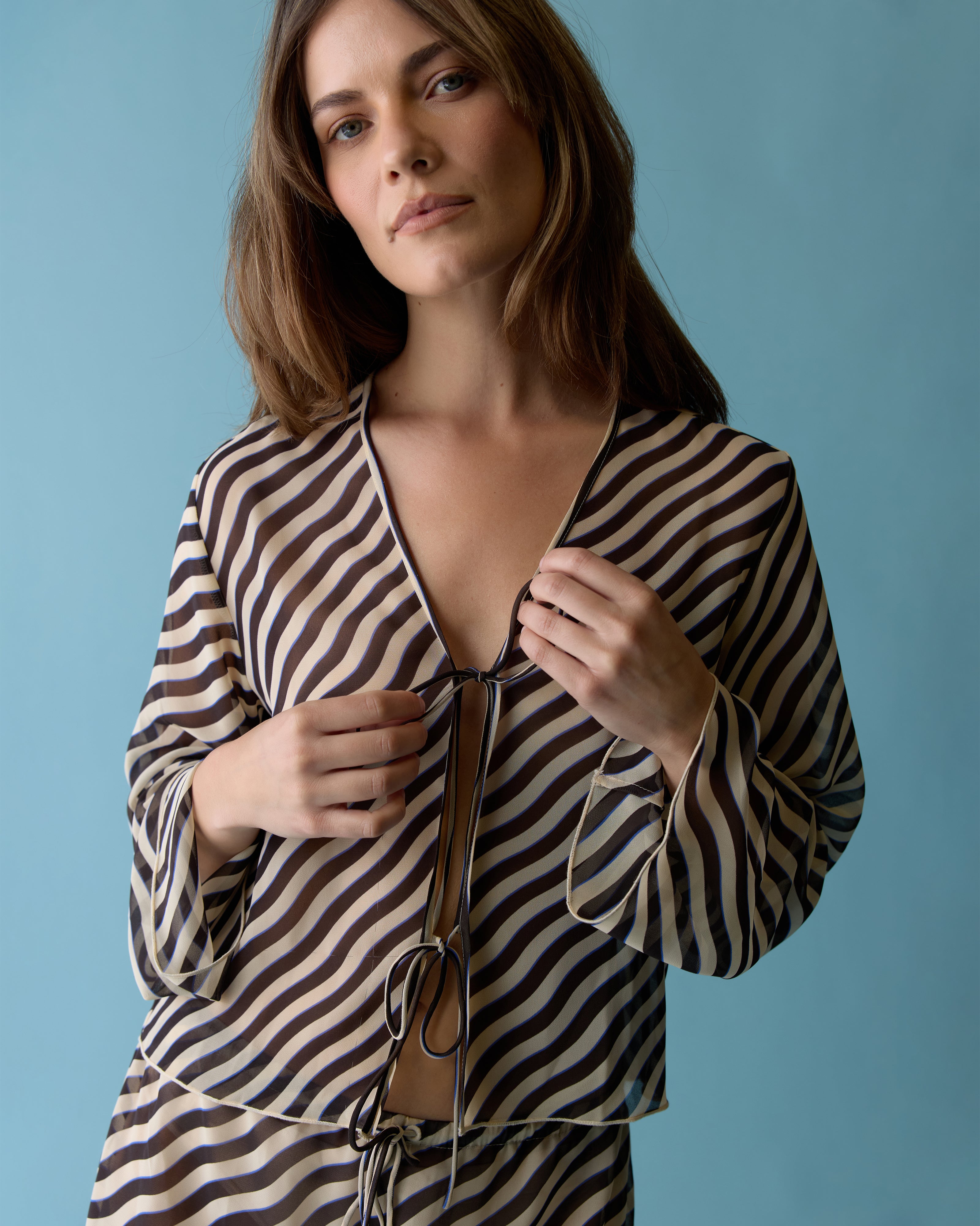 A woman stands against a blue background, wearing The Curacao Top - Tierra by Dandy Del Mar, featuring striped fabric, long bell sleeves, and a tie front, looking directly at the camera.
