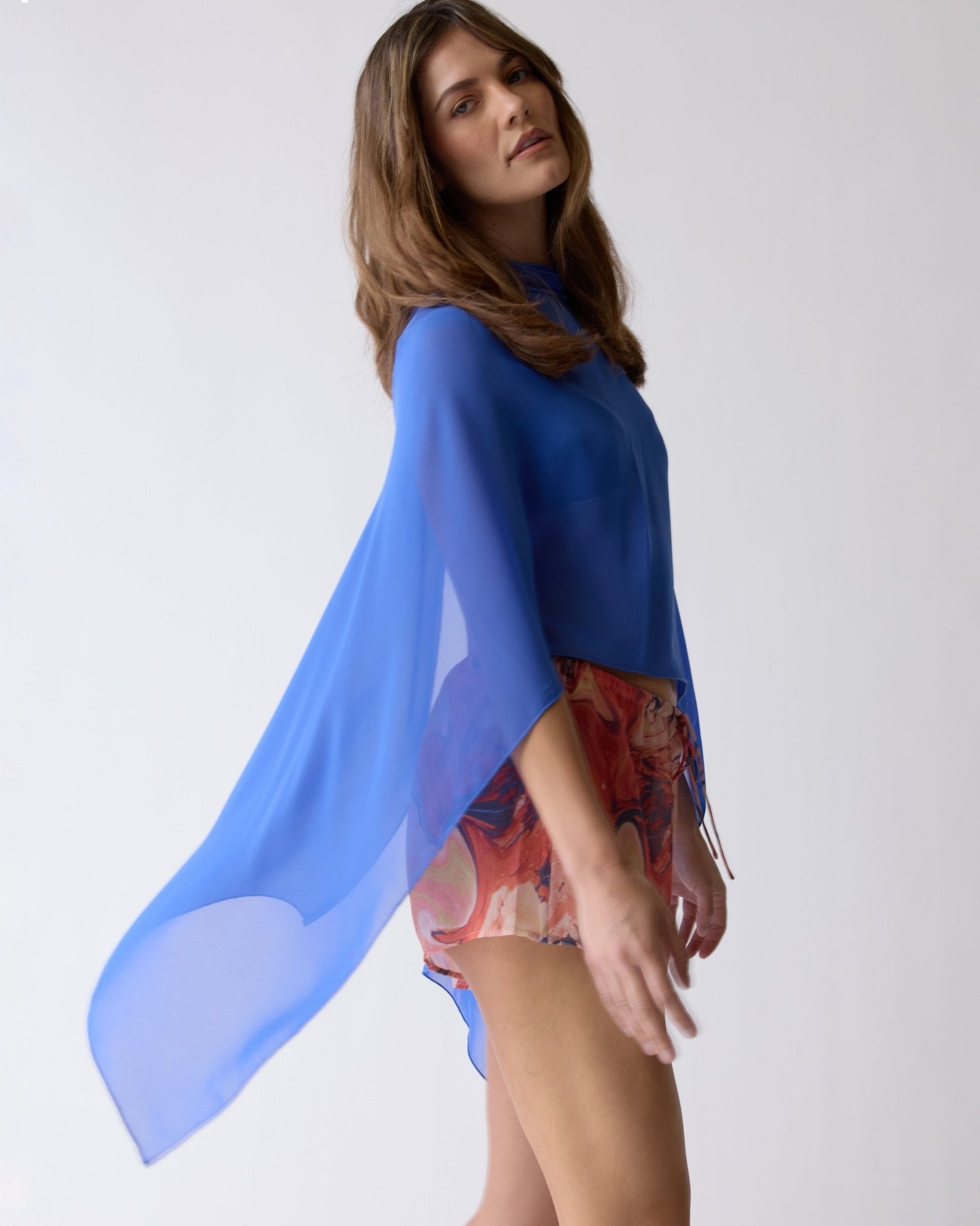 A woman poses against a plain background in The Zante Top - Solana by Dandy Del Mar, featuring an open back, sheer blue chiffon, and flowing sleeves—an effortlessly versatile look.