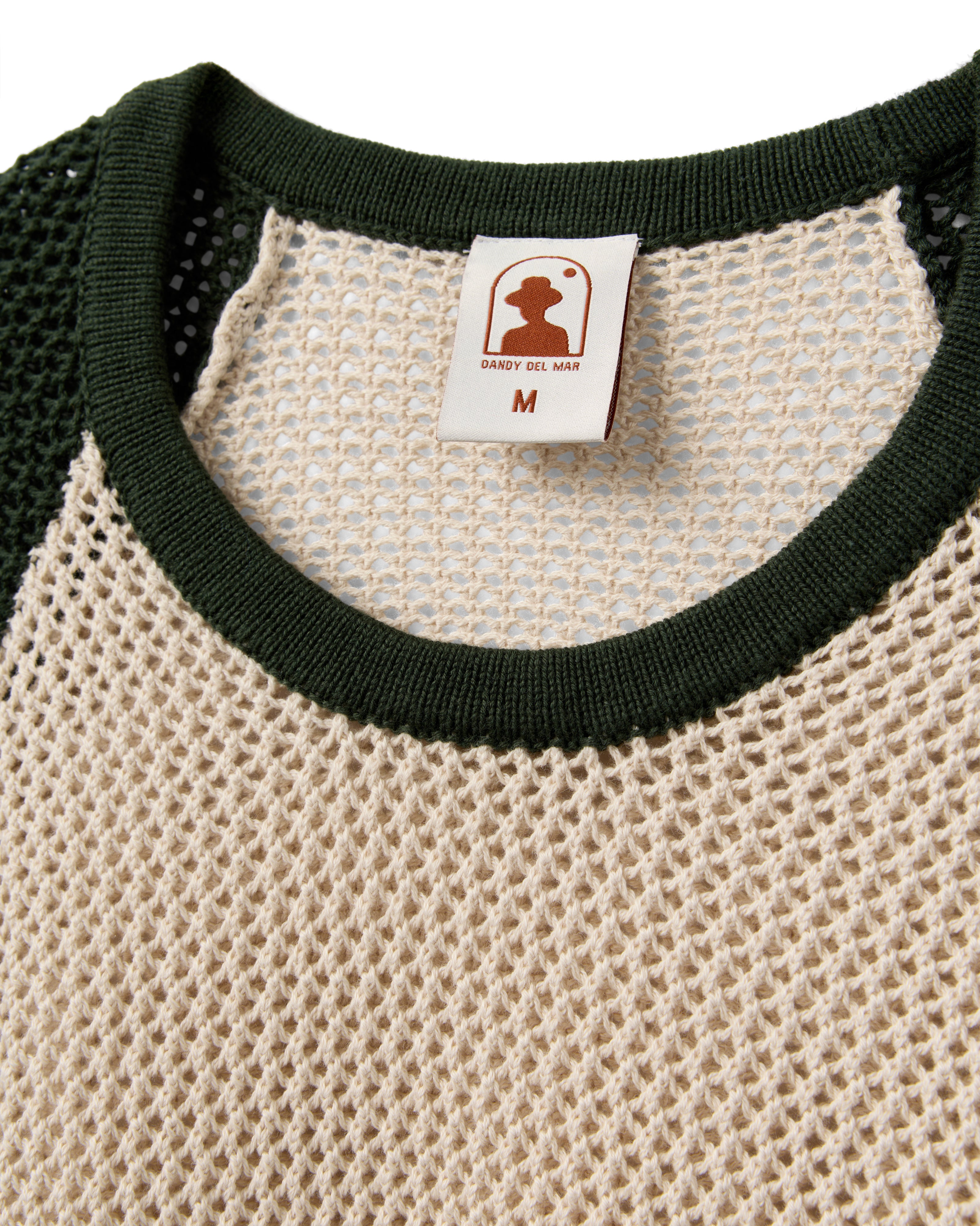Close-up of The Porto Crochet Tank - Sabbia by Dandy Del Mar, a beige open-knit mesh tank with green collar trim and a brand tag in size M.