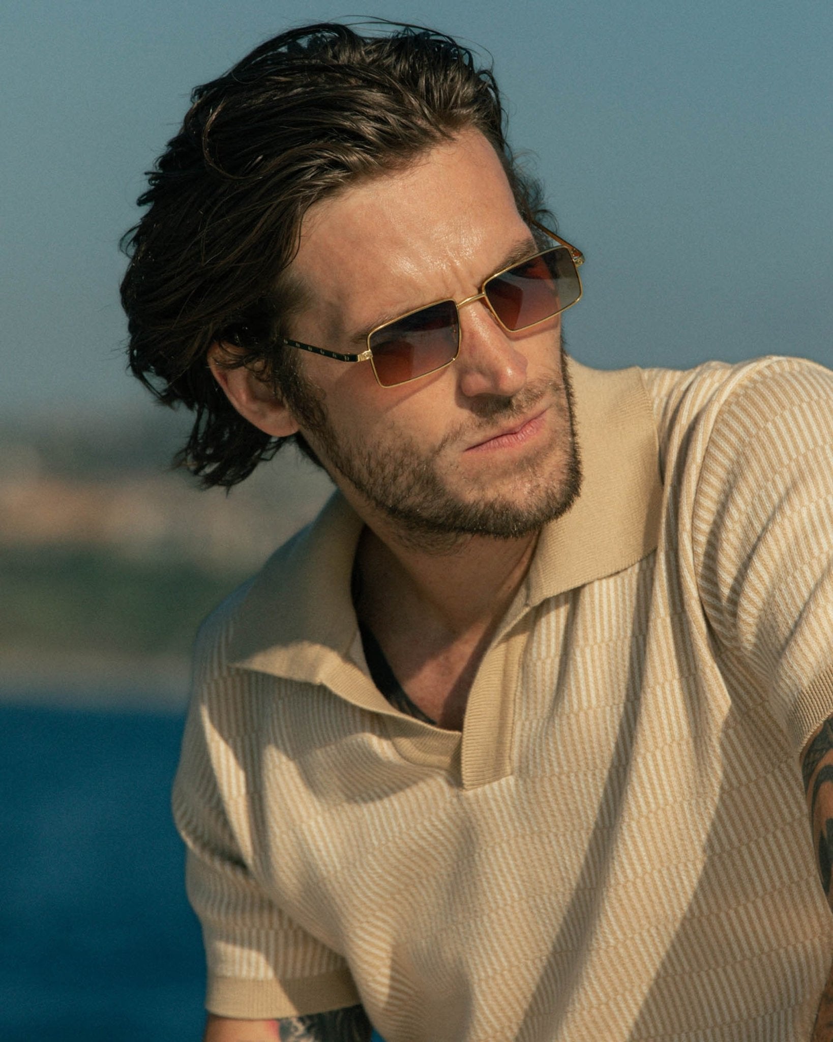 A man with shoulder-length hair and a beard wears sunglasses and the Dandy Del Mar Sebastian Knit Polo Shirt - Shell, glancing away from the camera with a scenic view of water and land in the background.