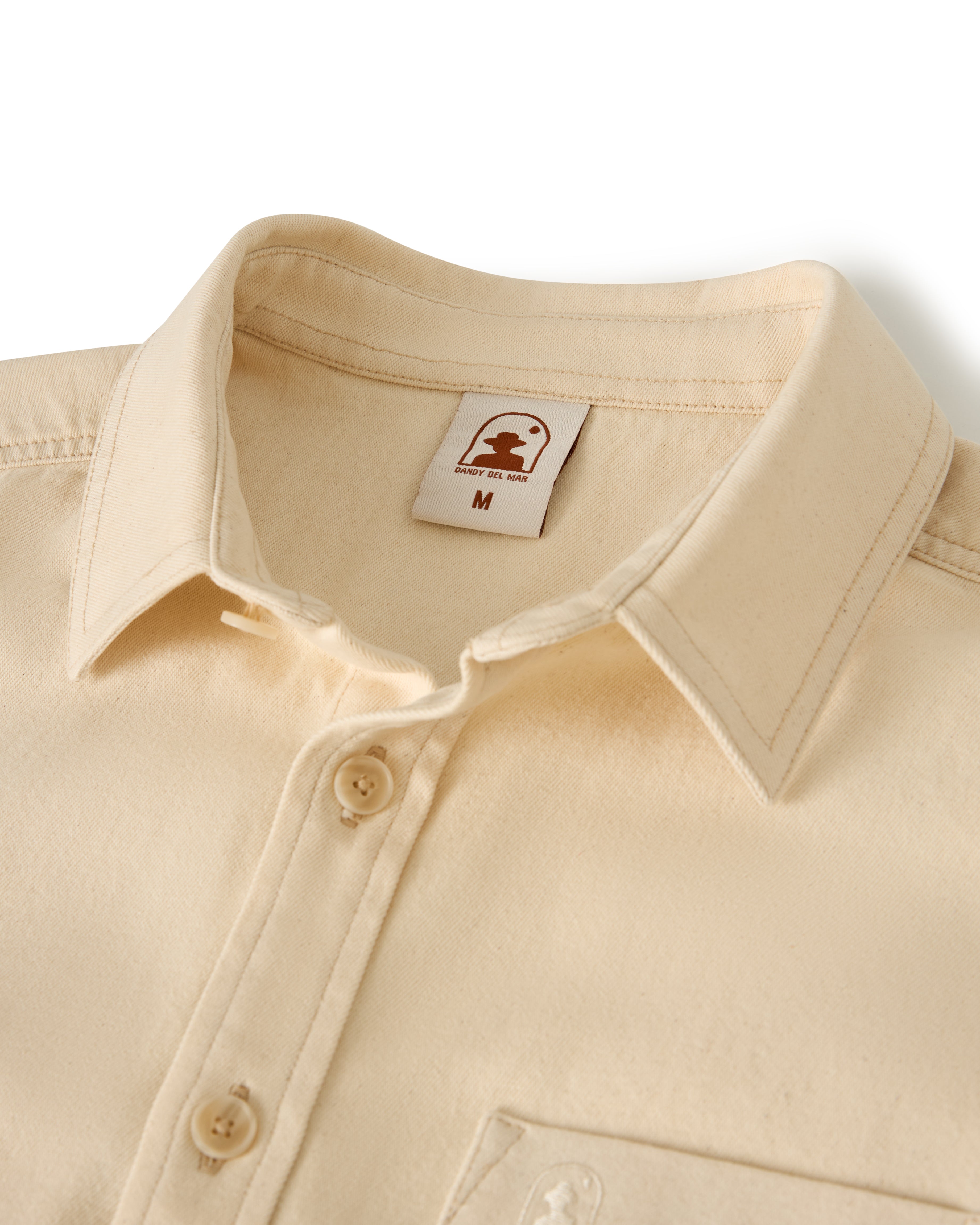 The Dandy Del Mar "The Cartagena Denim Shirt - Scallop" is shown. It's a beige button-up with a collar and features a label with an icon and "M" for medium size.