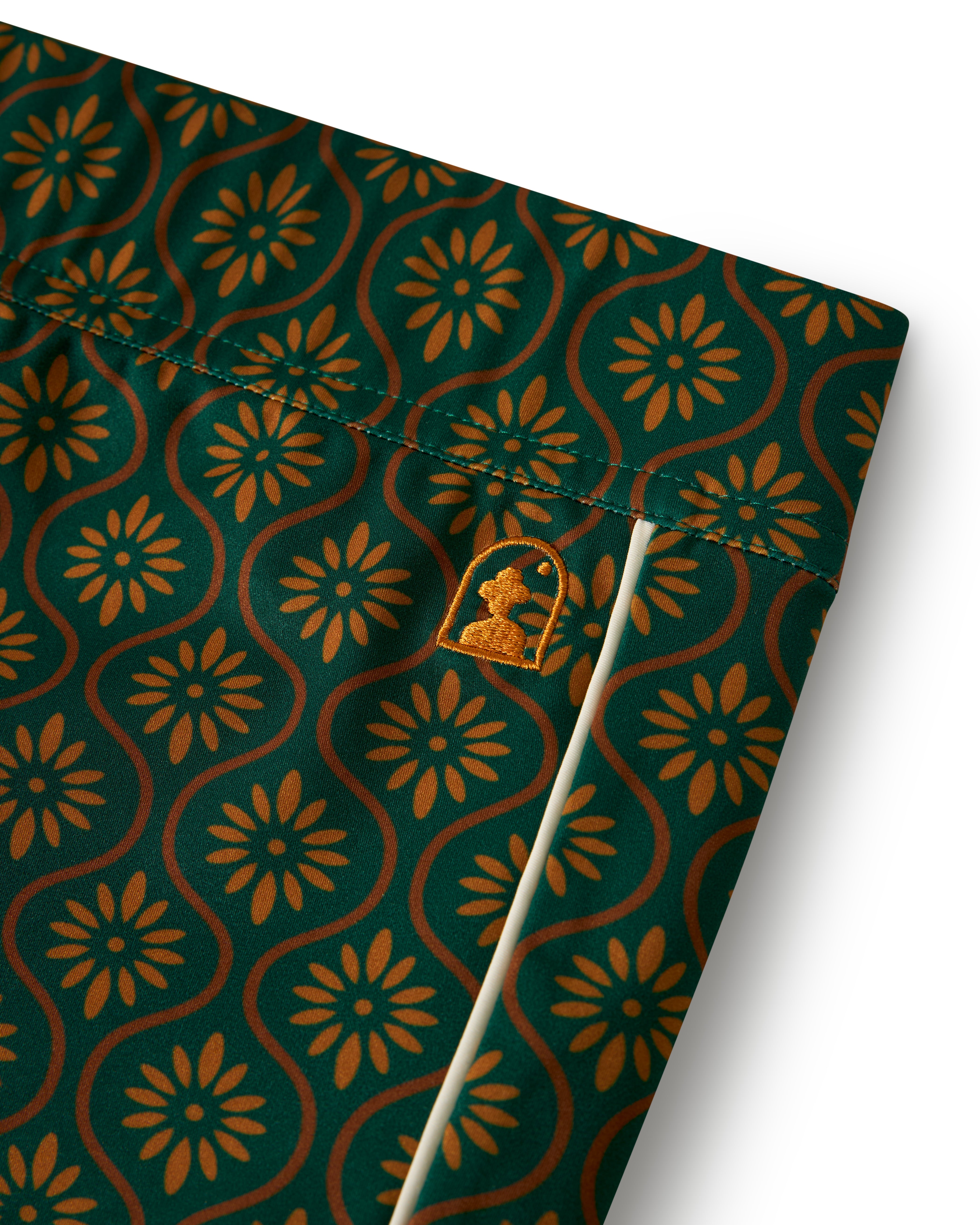 Close-up of The Cassis Square Cut Swim Brief - Albero by Dandy Del Mar, featuring a green and orange floral pattern on European fit swim briefs. A small embroidered logo of a stylized silhouette is visible near the seam, crafted from recycled polyester.