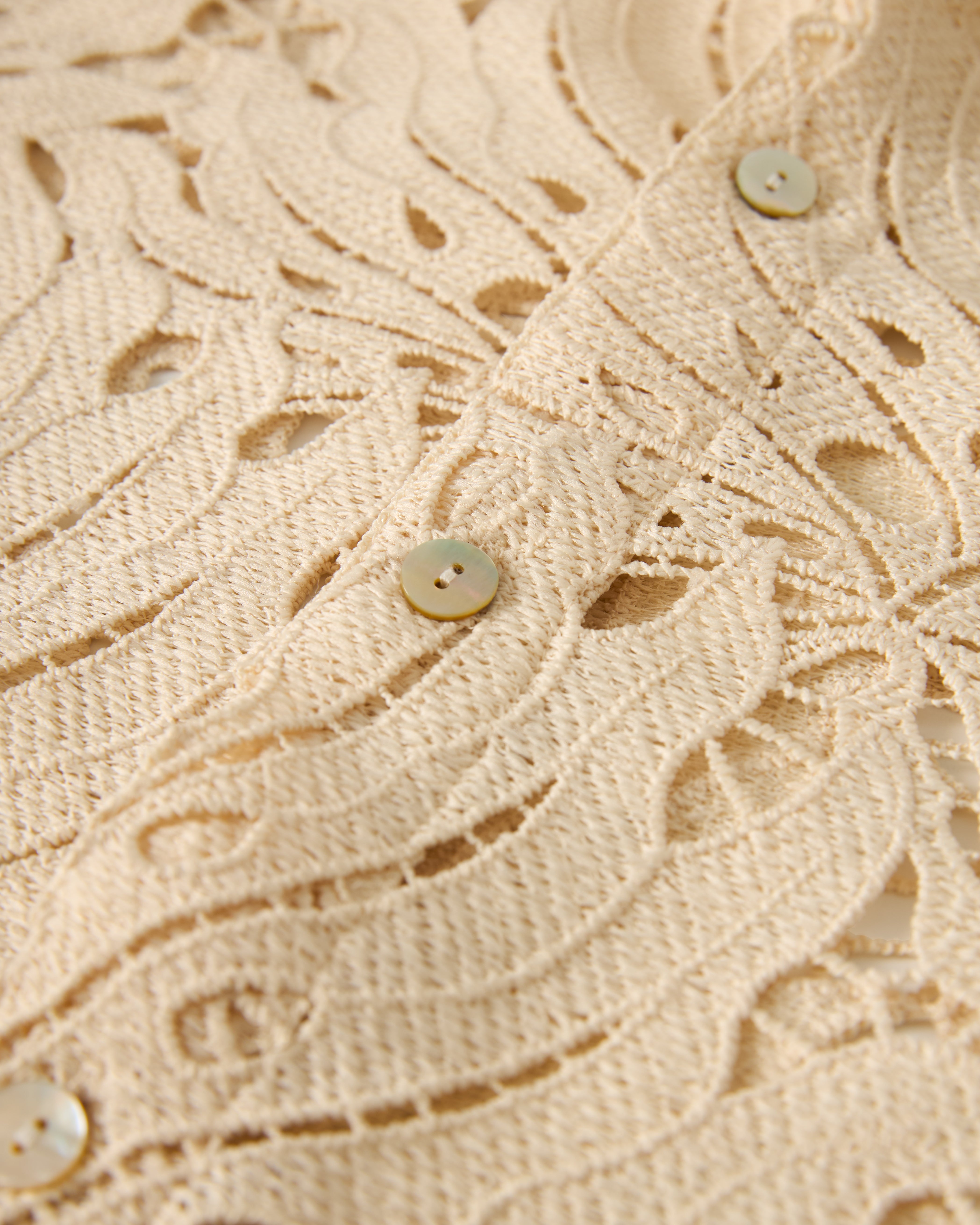 Close-up of The Monstera Shirt - Scallop by Dandy Del Mar, showcasing beige lace fabric with a floral pattern and round buttons.