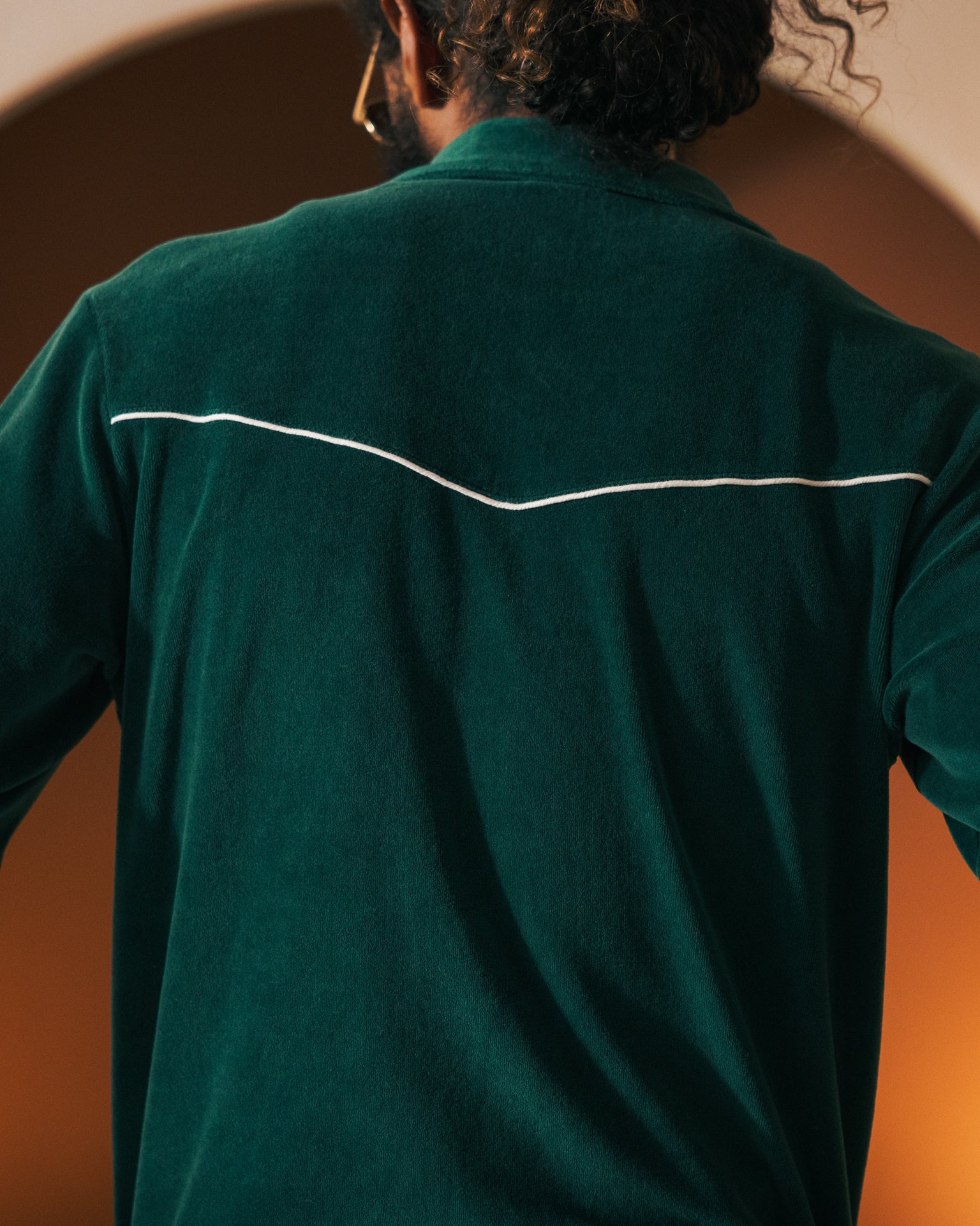 A person with curly hair is seen from behind, wearing The Rio Velour Jacket - Albero by Dandy Del Mar, a dark green jacket featuring a classic track silhouette and a thin white line across the upper back.
