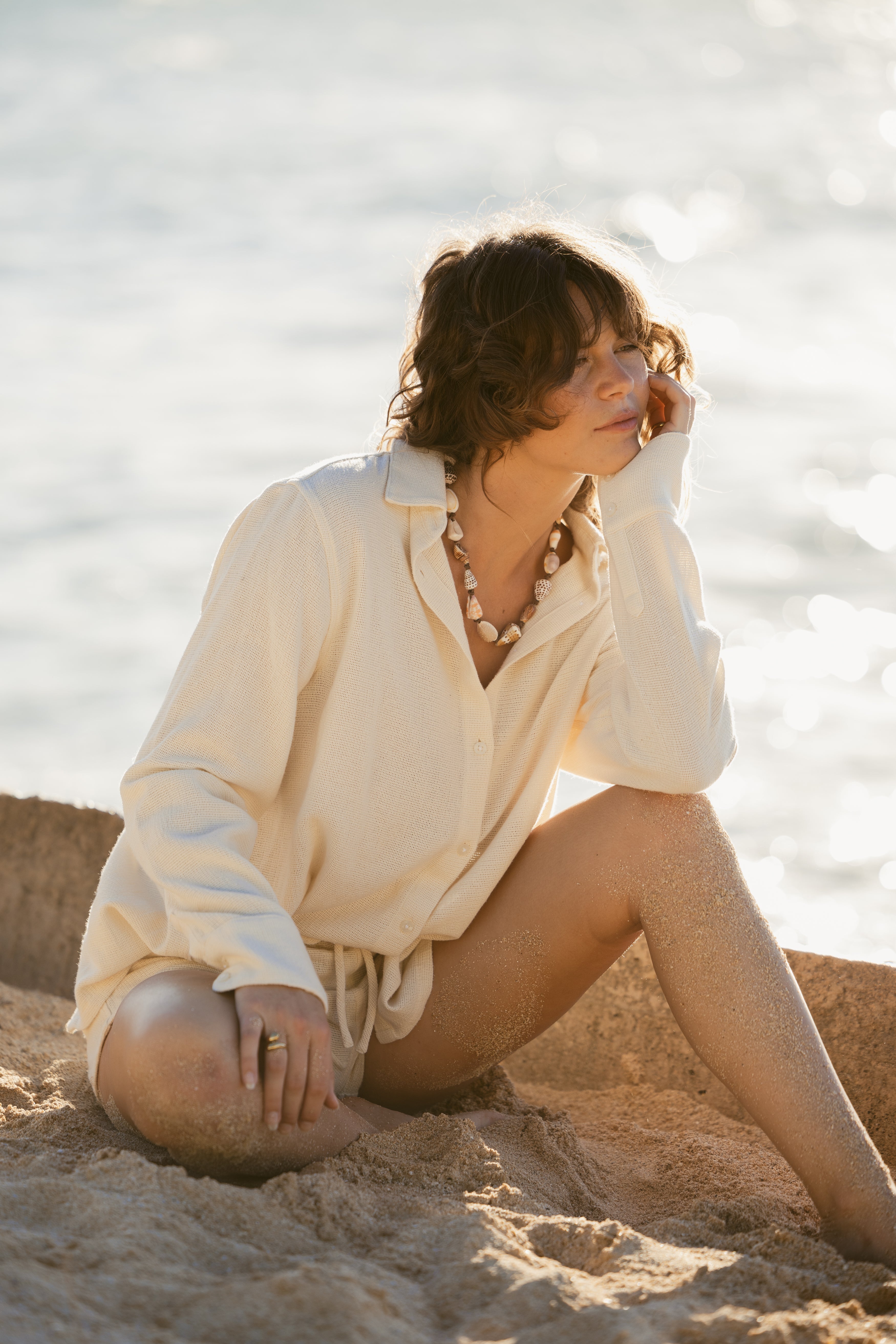 Wearing the Dandy Del Mar Sevilla Top - Scallop and a shell necklace, a person sits on the sandy beach with one hand on their face, gazing out at the water.