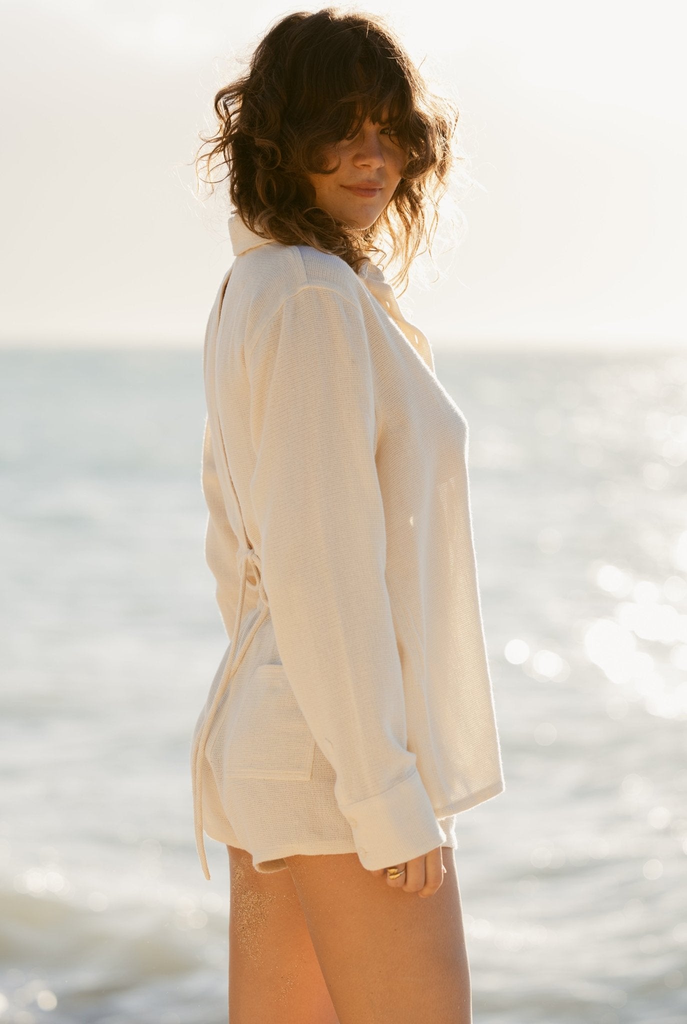 On the beach with the ocean behind them, a person with wavy hair wears a long-sleeve white shirt and Dandy Del Mar’s The Sevilla Shorts - Scallop, featuring an elastic waistband, standing partially turned from the camera.