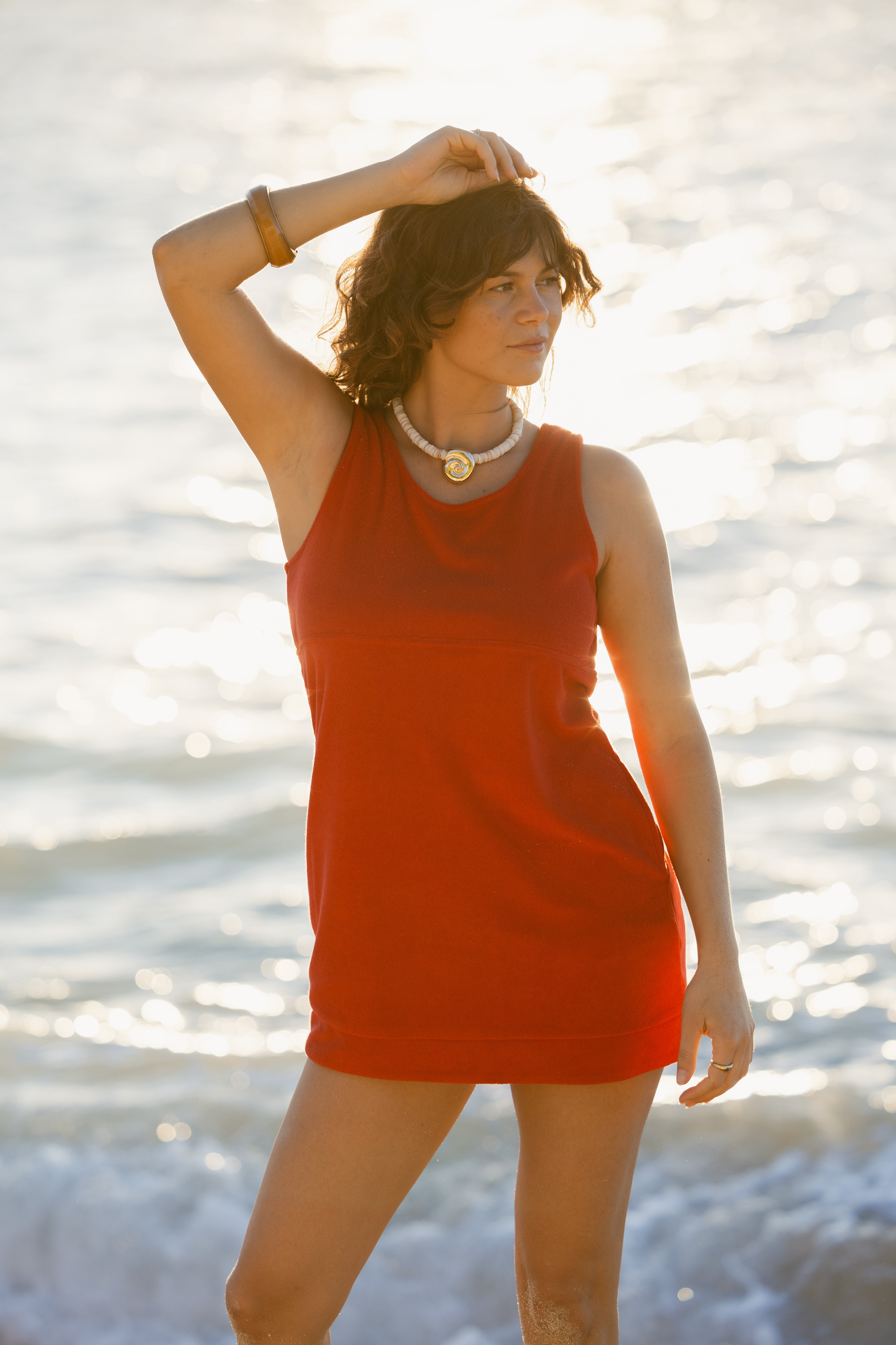 Wearing The Cyprus Dress - Pico by Dandy Del Mar, a person stands on a sunny beach with shimmering water, one arm raised while the other rests by their side.