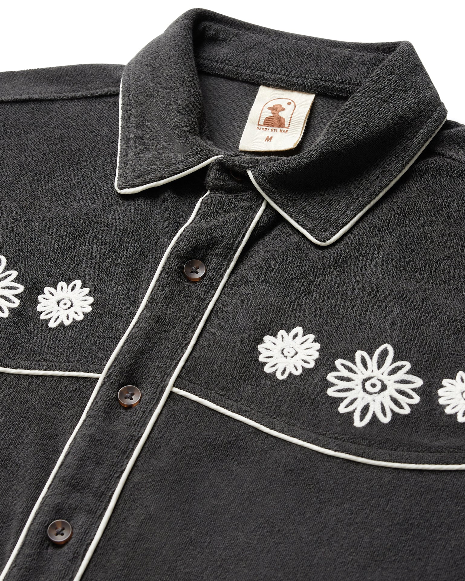 A close-up of Dandy Del Mar's The Gaucho Shirt - Onyx reveals white floral embroidery and contrasting trim on black fabric. Brown buttons add a touch of equatorial cowboy flair.