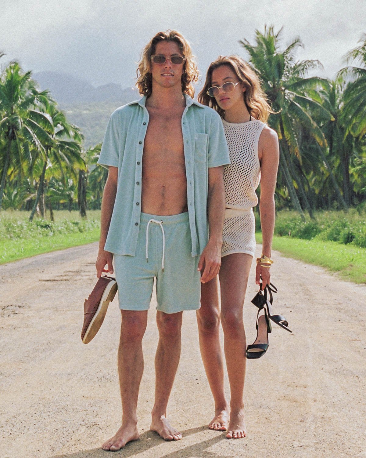 A barefoot man in Dandy Del Mar’s The Tropez Terry Cloth Shorts in Gulf Blue stands next to a barefoot woman holding black sandals, both on a palm-lined dirt road.