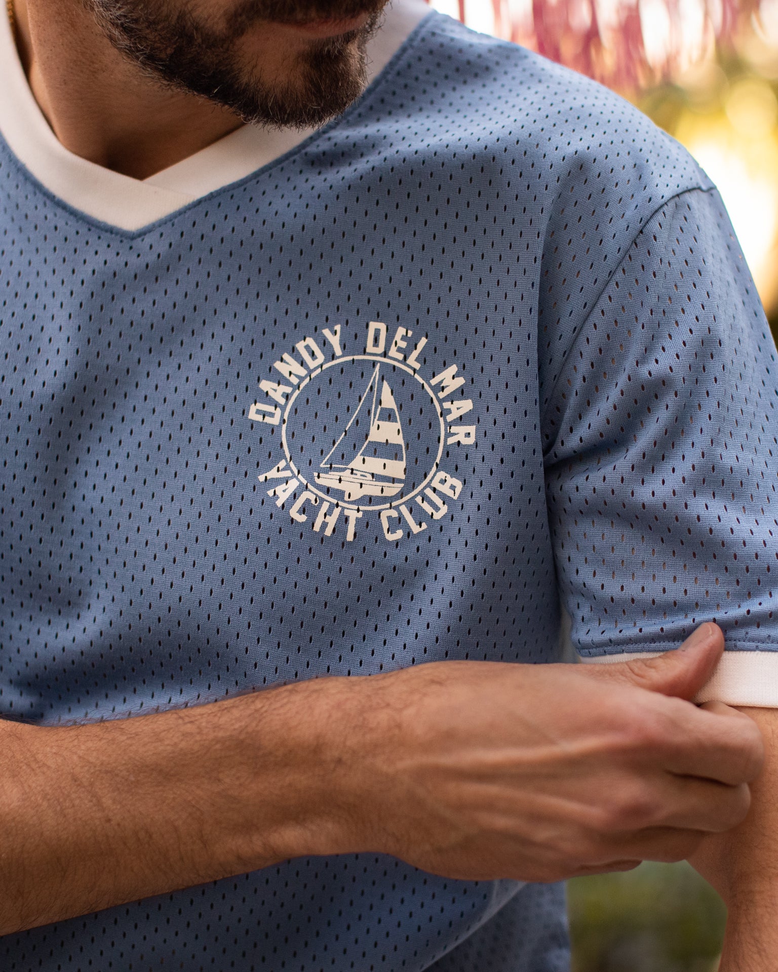 A person wearing The Kaena Mesh Tee - Annapolis from Dandy Del Mar exudes casual elegance with its blue sports mesh design featuring white trim and a sailboat graphic that reads "Candy Del Mar Yacht Club." The themed screen print beautifully complements the V-neck ribbing, creating an effortlessly stylish look.