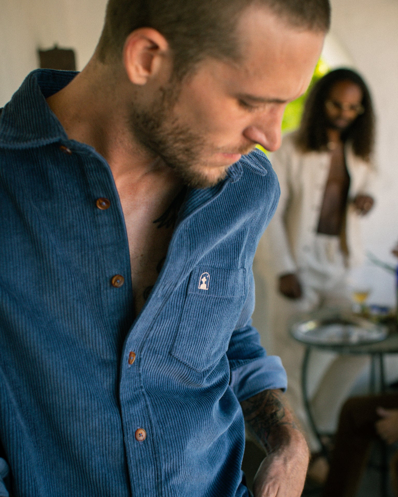 A man in a Moontide Corsica Corduroy Shirt from Dandy Del Mar looks down, while another person in the background dressed in white stands near a table with drinks.