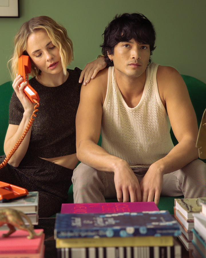 A woman wearing the Dandy Del Mar Dominica Crochet Tank in Vintage Ivory talks on a vintage orange telephone, sitting close to a man who appears pensive, in an environment with retro aesthetics.