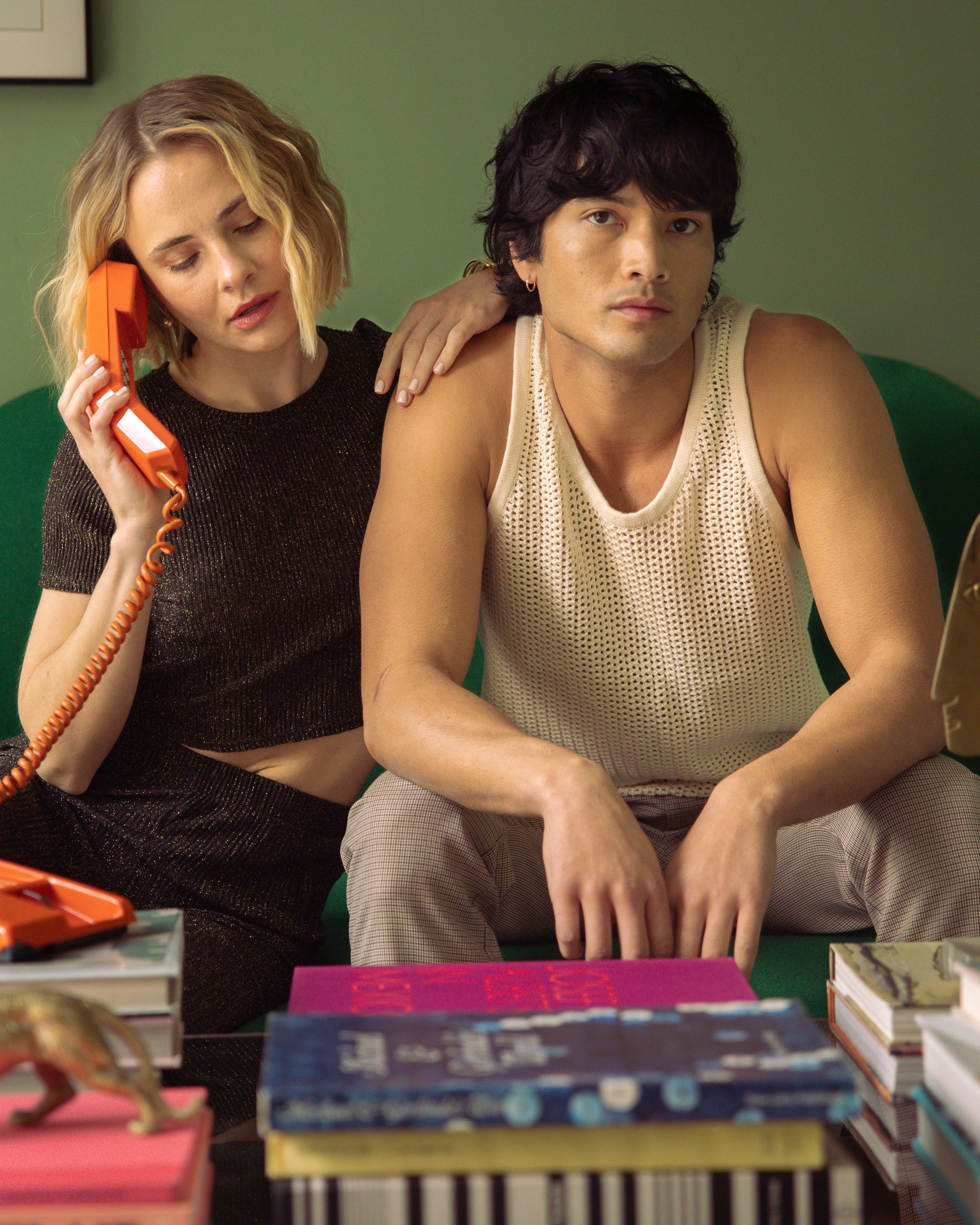 A woman wearing the Dandy Del Mar Dominica Crochet Tank in Vintage Ivory talks on a vintage orange telephone, sitting close to a man who appears pensive, in an environment with retro aesthetics.