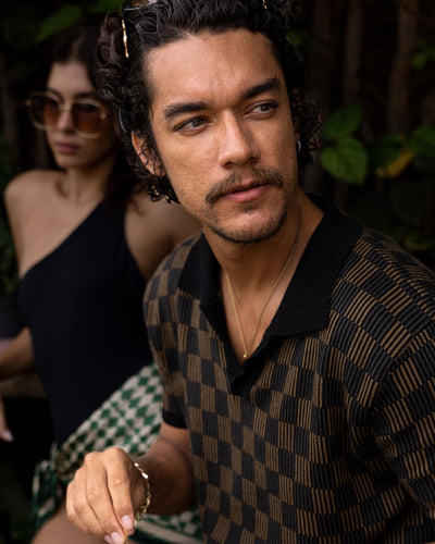 A man with curly hair and a mustache wears the Dandy Del Mar Sebastian Knit Polo Shirt in Onyx and gold jewelry; behind him, a woman in sunglasses and a one-shoulder top stands stylishly.
