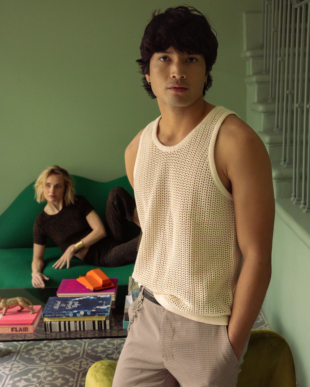 A young man in The Dominica Crochet Tank - Vintage Ivory by Dandy Del Mar stands in the foreground, while a woman sitting on a green couch looks on from the background, in a room with books on the coffee table.