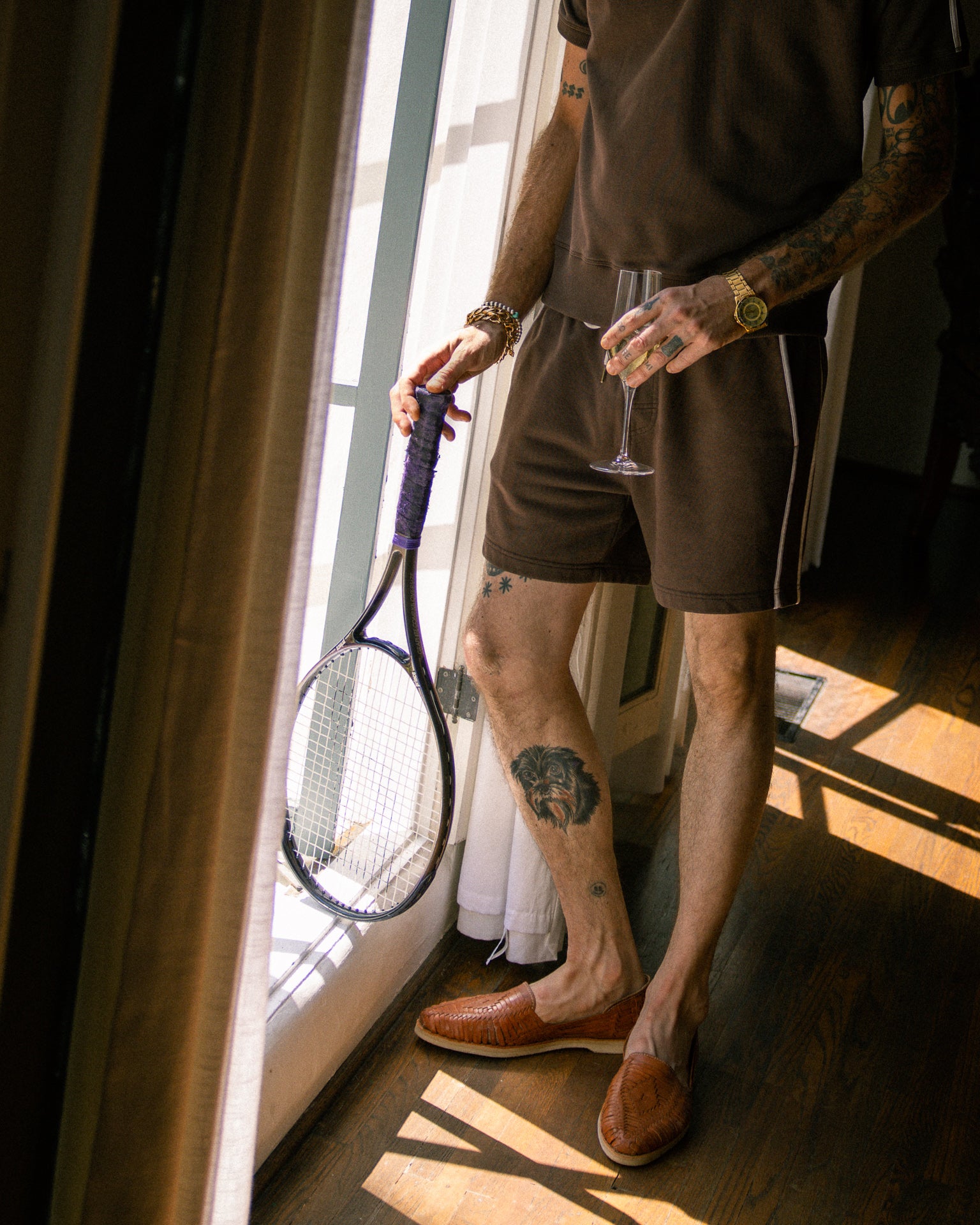 A person stands indoors wearing the Marseille French Terry Short - Carajillo by Dandy Del Mar, holding a tennis racket in one hand and a wine glass in the other. One leg shows a large tattoo. The individual is wearing brown slippers.