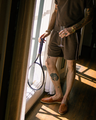 Wearing the Dandy Del Mar Marseille French Terry Shorts in Carajillo, a person with visible arm and leg tattoos stands by a window in casual attire, holding a tennis racket and wine glass, paired with brown slip-on shoes.