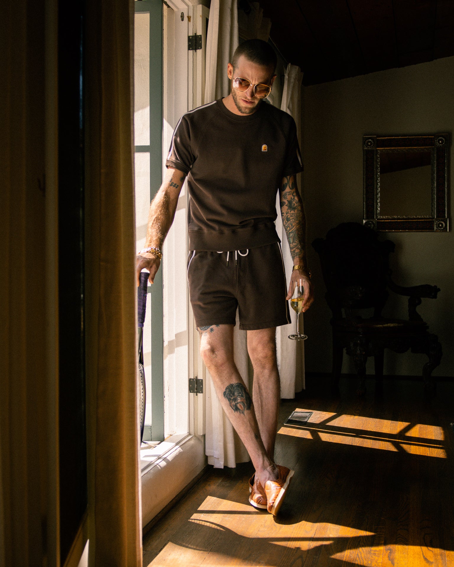 A tattooed individual, donning sunglasses and a dark brown Marseille French Terry Pullover - Carajillo by Dandy Del Mar with a ribbed neckband, stands by a window holding a glass of wine and a tennis racket.