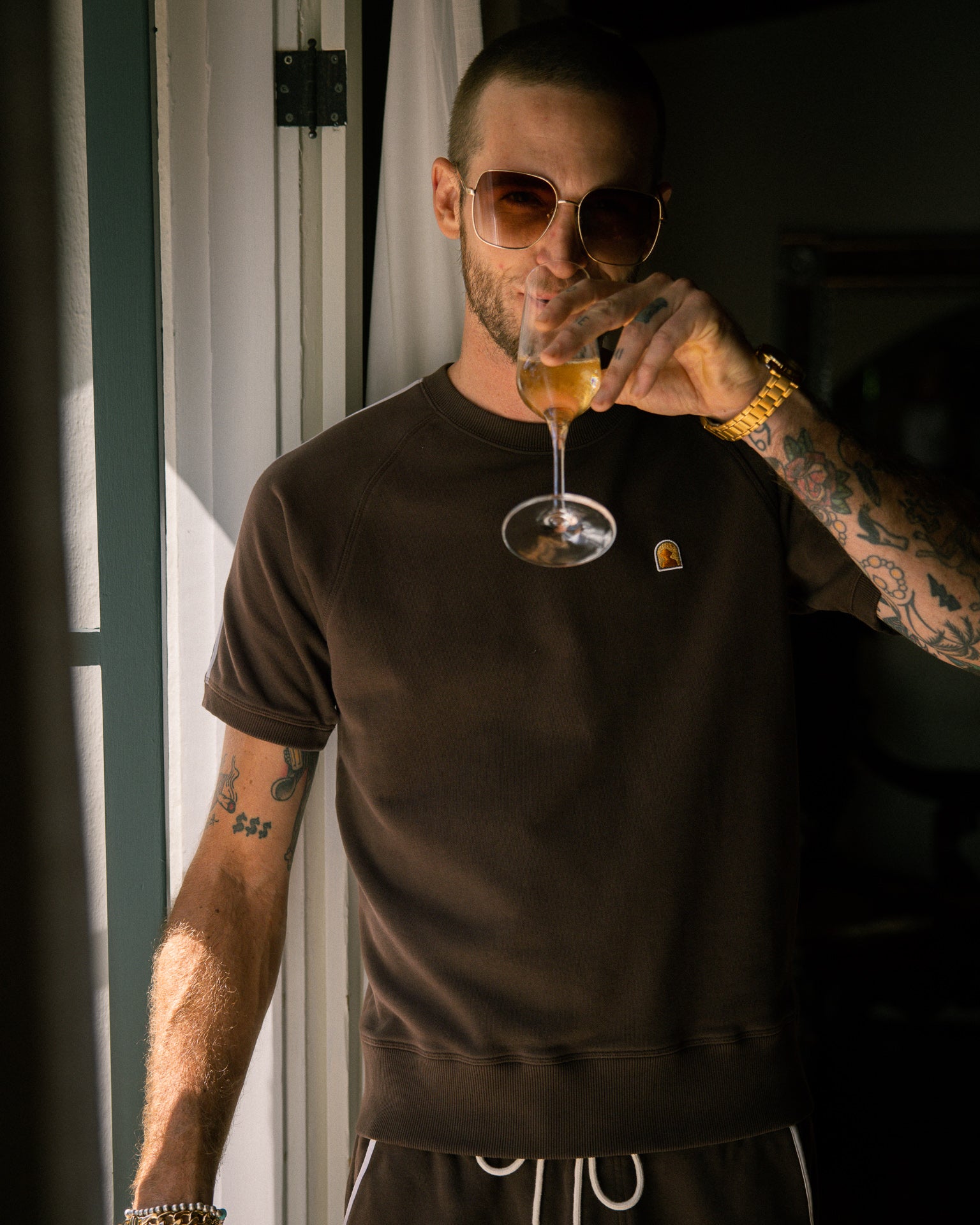 A man wearing The Marseille French Terry Pullover - Carajillo by Dandy Del Mar, adorned with tattoos and sunglasses, sips a drink from a wine glass while standing by a doorway.