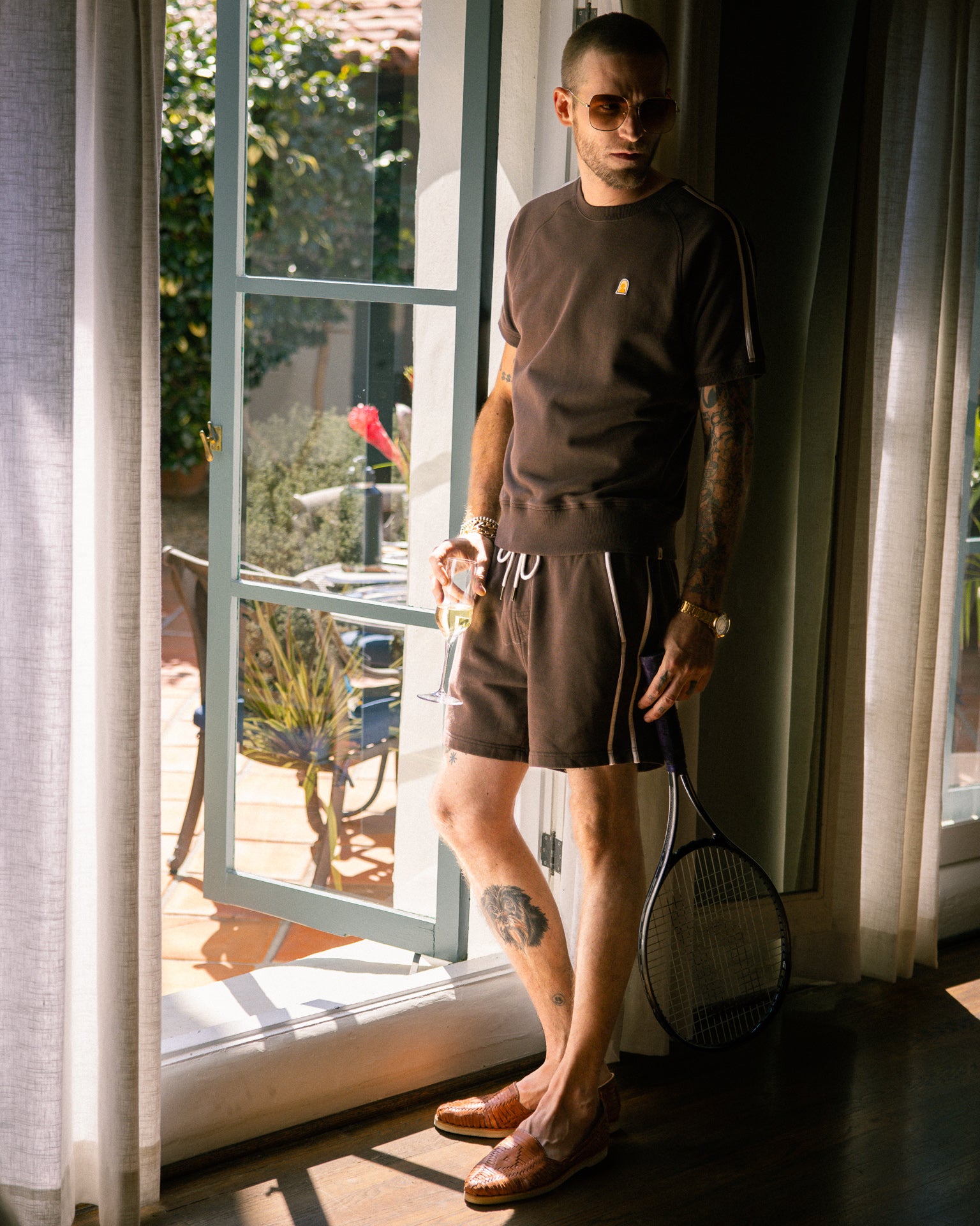 A man wearing sunglasses, a matching brown shirt, The Marseille French Terry Short - Carajillo by Dandy Del Mar, and brown shoes stands indoors near an open door, holding a tennis racket.