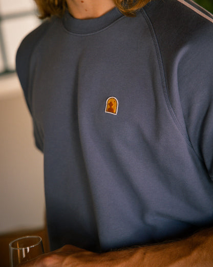 Person wearing The Marseille French Terry Pullover in Moontide by Dandy Del Mar, holding a glass, with the image focusing on the upper body and featuring an embroidered patch on the chest.