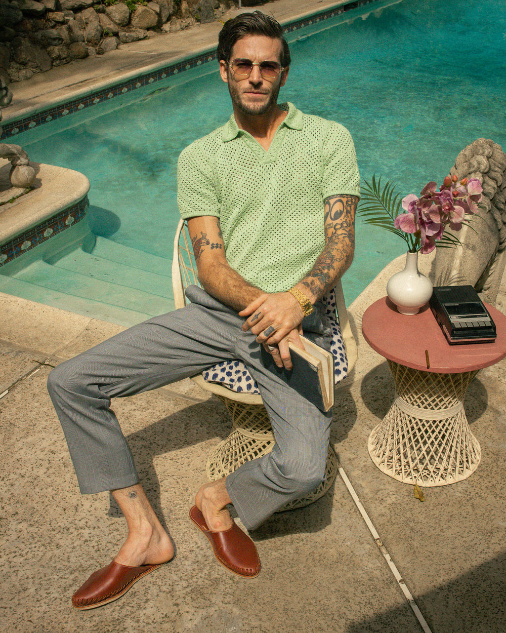 A man in a Dandy Del Mar Antibes Crochet Shirt - Pistachio and trousers sitting casually by a pool with sunglasses on.