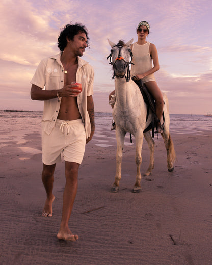 Amid a pink and purple sky evoking the serene Equatorial Cowboy vibe of dawn, someone strolls barefoot on the beach in The Gaucho Shorts - Alabaster by Dandy Del Mar, holding an apple as another rides a white horse beside them.