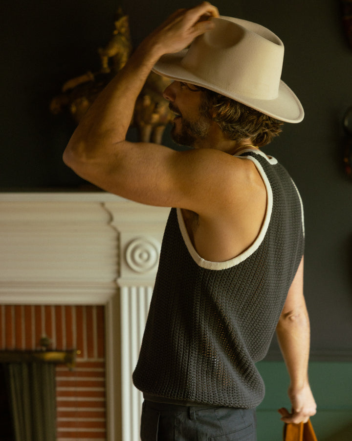 Man in a Dominica Crochet Tank by Dandy Del Mar tipping his hat in a room with a fireplace.