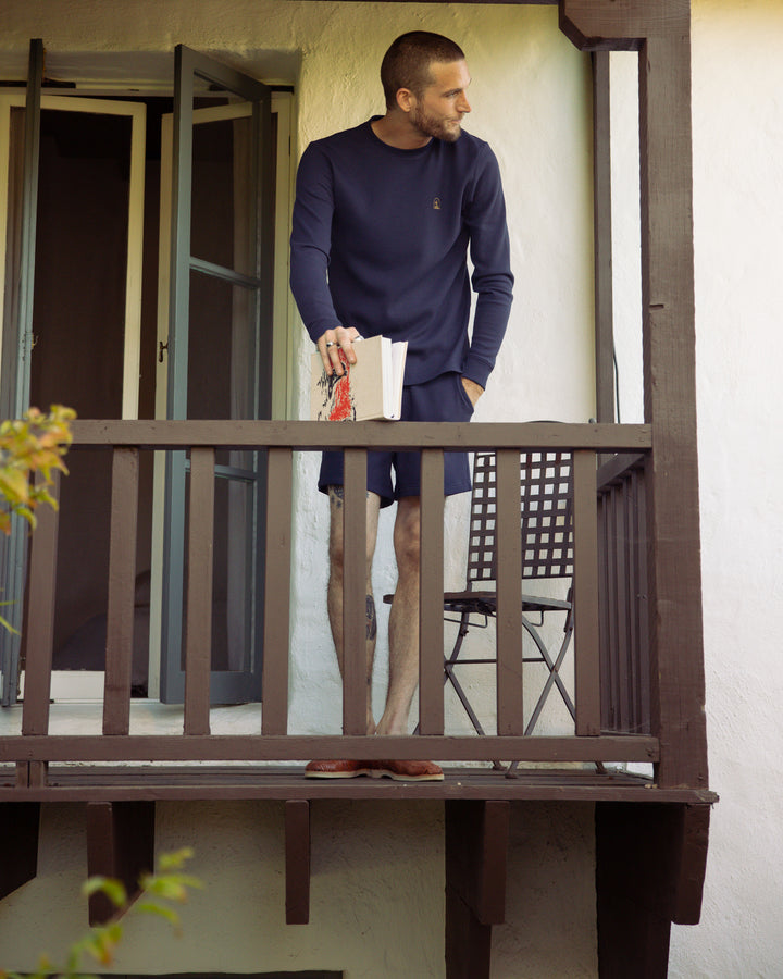 A man standing on a balcony holding a book wearing The Cannes Short - Luxe Navy by Dandy Del Mar.