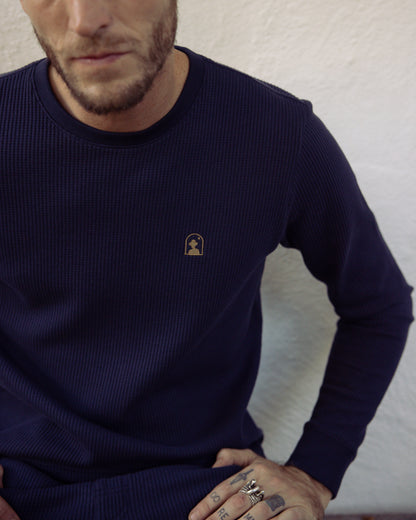 A person wearing a Luxe Navy Cannes Long Sleeve Shirt by Dandy Del Mar, featuring an embroidered logo on the chest, and several rings on their fingers sits against a white background.