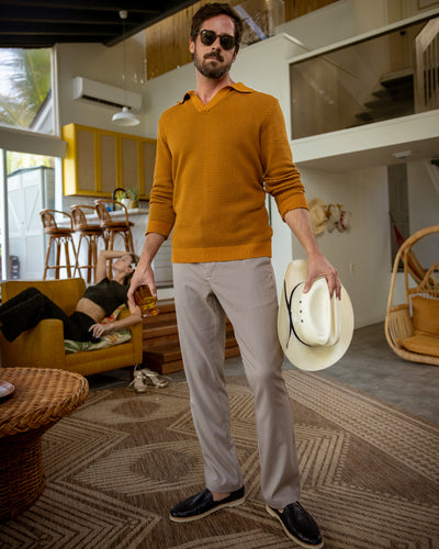 A man in Dandy Del Mar's The Sebastian LS Polo Shirt in Burnt Sienna, beige pants, and black shoes stands indoors with sunglasses, holding a white cowboy hat and a drink; a woman lounges on a green couch in the background.