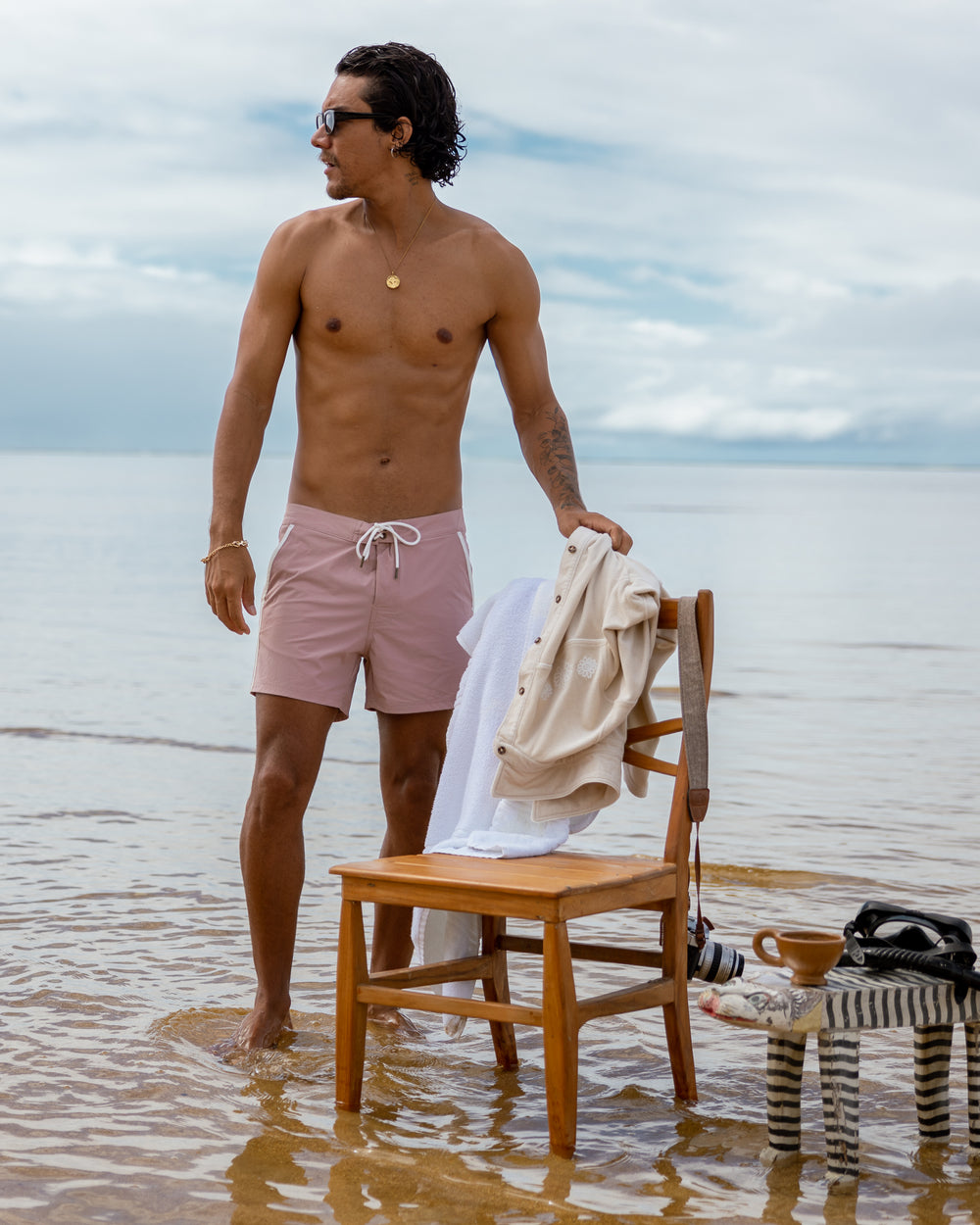 A man in pink swim trunks standing next to a chair in the water. The Stirata Swim Short - Rosado by Dandy Del Mar is made from performance fabric, providing 4-way stretch for ultimate comfort.