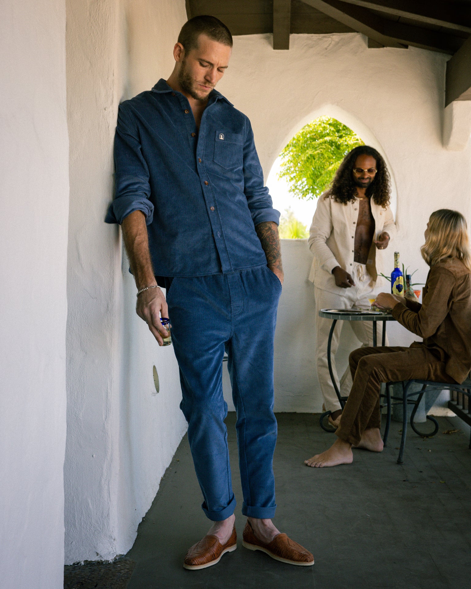 Three men are depicted in a casual clothing setting: one leans against a white wall wearing The Corsica Corduroy Pant - Moontide by Dandy Del Mar, another relaxes at a table, and the third man stands nearby dressed in 8-Wale corduroy. They're all dressed in relaxed, comfortable attire.