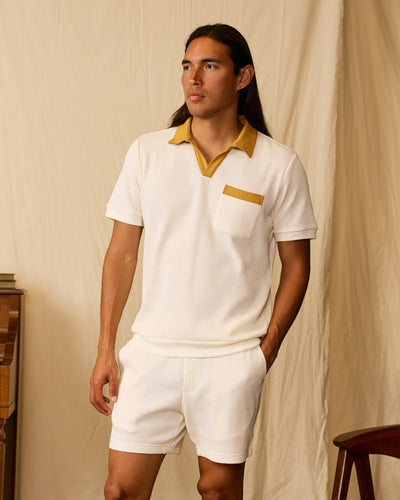 A man wears Dandy Del Mar’s The Cannes Waffle Knit Polo Shirt in Vintage Ivory— a buttonless polo with a mustard collar and pocket detail—paired with white shorts, standing indoors against a beige backdrop.
